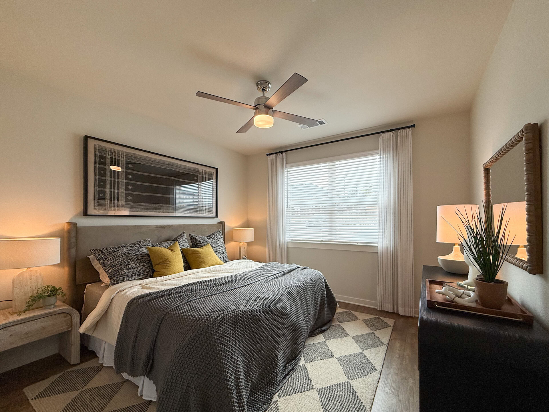 A cozy, spacious bedroom features a queen-sized bed. A modern ceiling fan with a light fixture hangs above. A large window with white blinds allows natural light to enter.