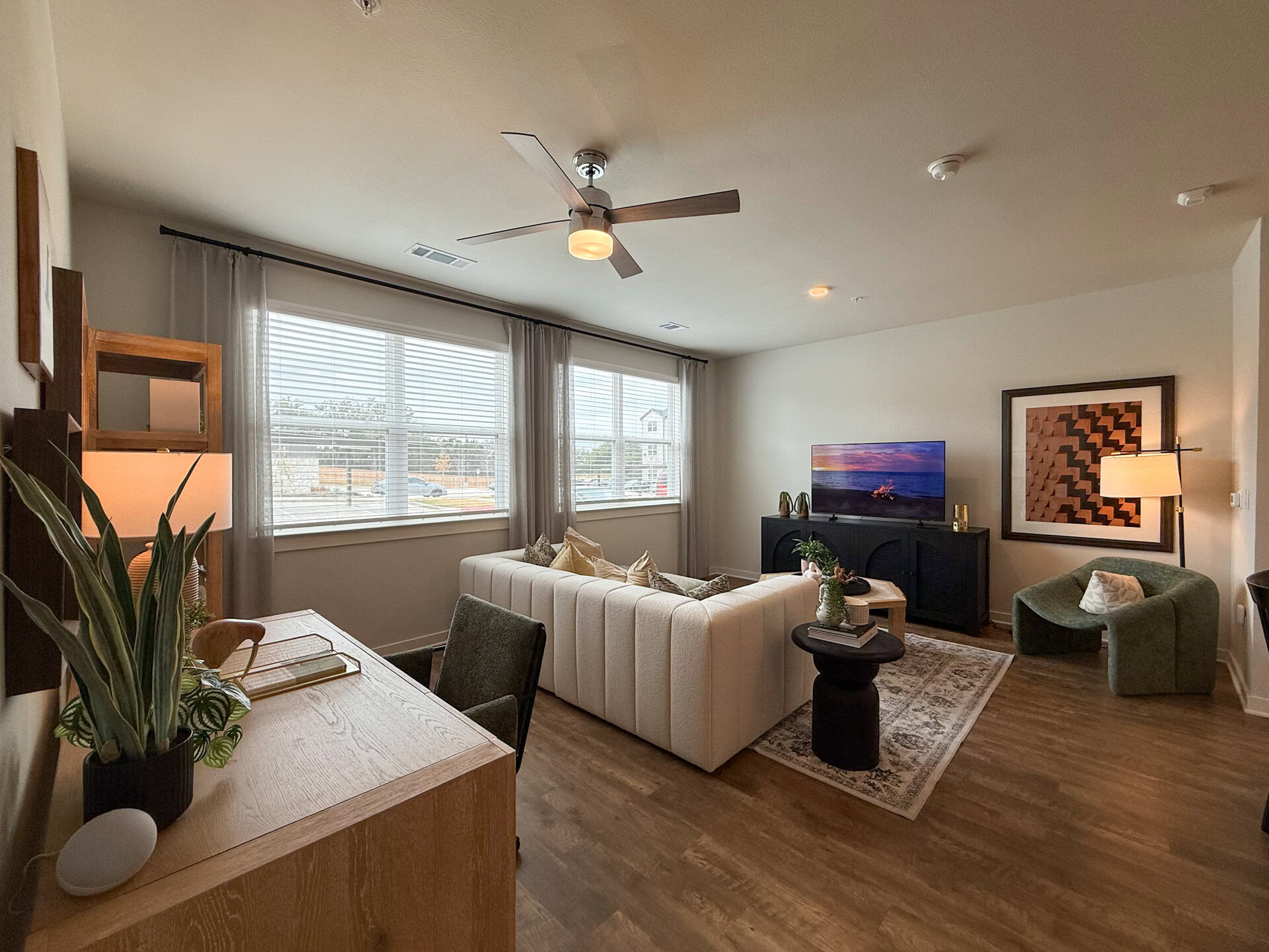 A spacious modern living room features Natural light that streams through large windows adorned with sheer curtains, illuminating the space. The flooring is a warm wood tone, enhancing the cozy atmosphere.