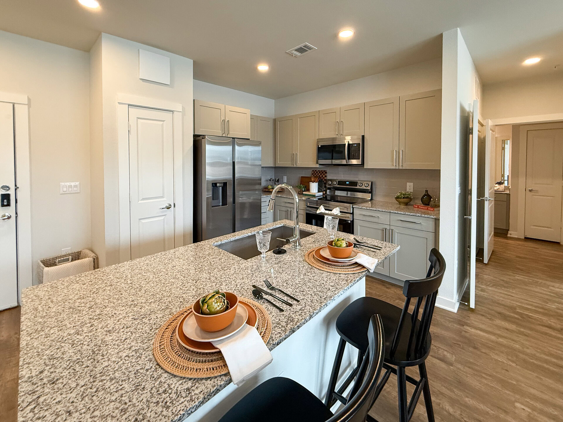 A modern kitchen features light-colored cabinetry and Samsung stainless steel appliances, including a refrigerator and oven. The countertop is made of speckled granite, with a sink integrated into the island. The space is well-lit with natural light and has a warm wooden floor.