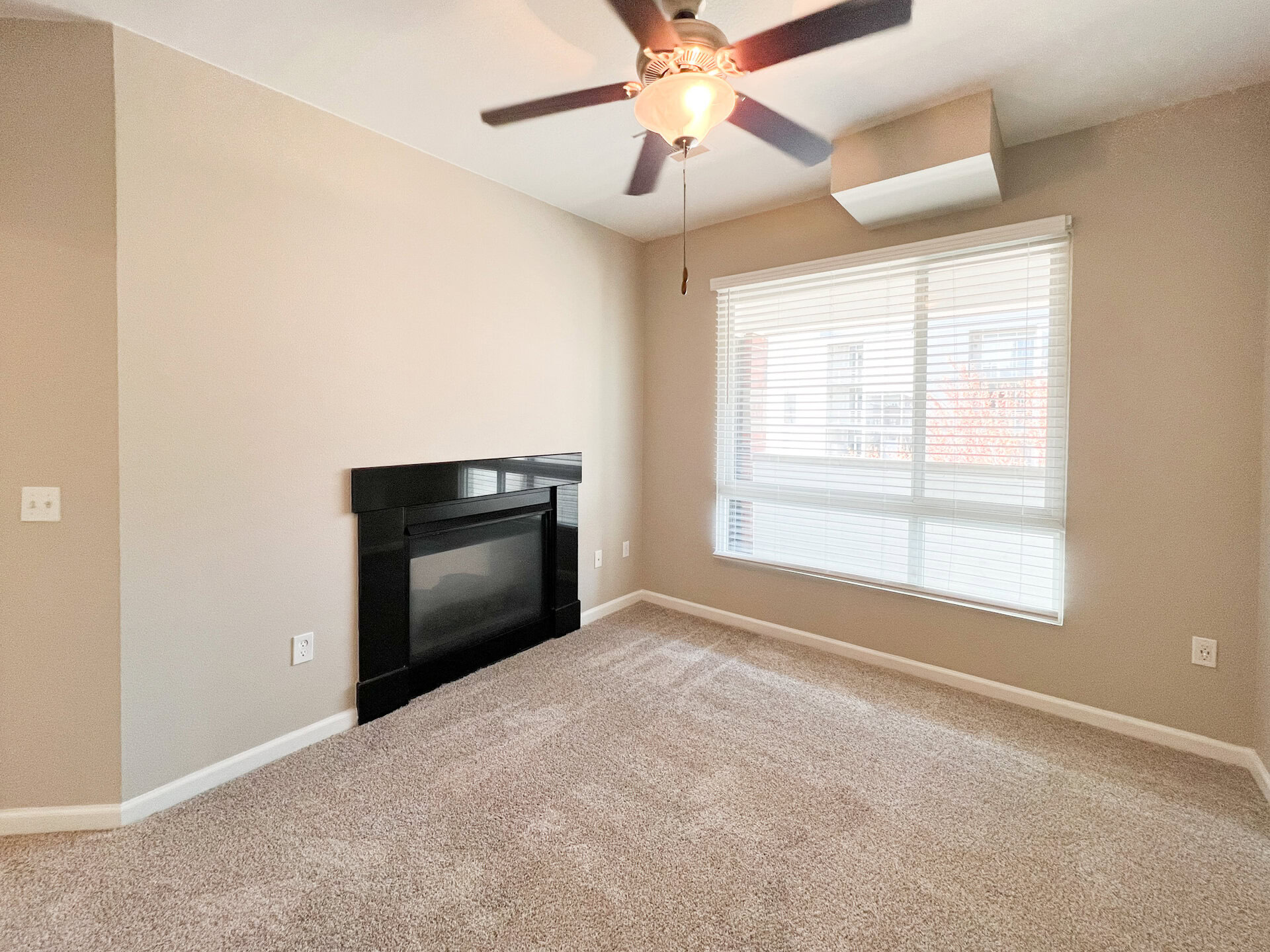 A cozy bedroom features light beige walls and plush gray carpeting. A sleek black fireplace is positioned against one wall, while a large window with white blinds allows natural light to fill the space. A ceiling fan hangs above, adding to the room's comfort. The overall atmosphere is inviting and well-lit.