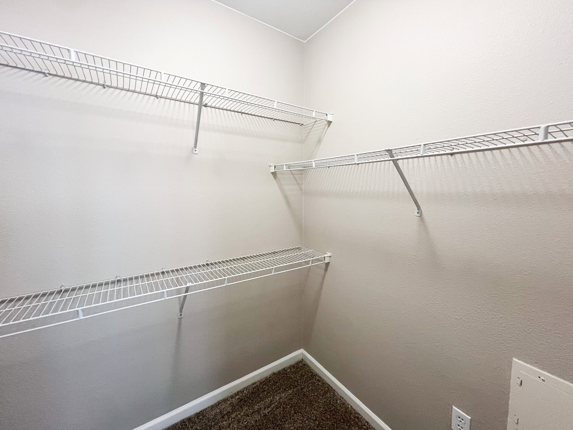A small closet features several white wire shelving units mounted on the walls. The shelves are arranged at different heights, providing ample storage space. The walls are painted a light gray, and the floor is carpeted in a neutral tone. The overall appearance is clean and organized, suitable for storing various items.