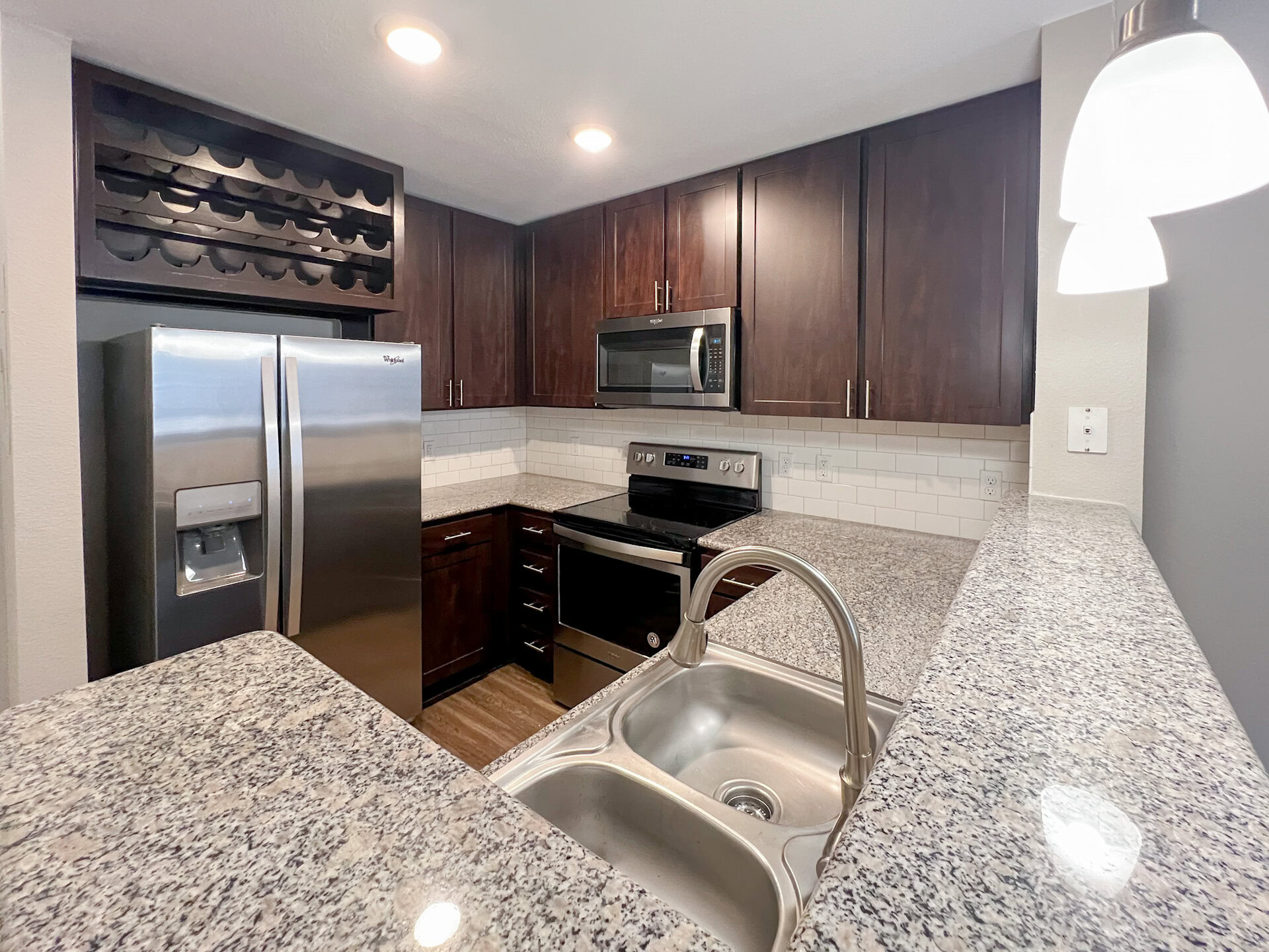 A modern kitchen featuring dark wood cabinetry and stainless steel appliances. The refrigerator is side-by-side with an ice and water dispenser. Below a microwave, there is a black electric stove with an oven. The countertops are made of speckled granite, and a double sink with a sleek faucet is visible. The backsplash consists of white subway tiles, and there is a wine rack above the refrigerator. The flooring appears to be a wood-like laminate.