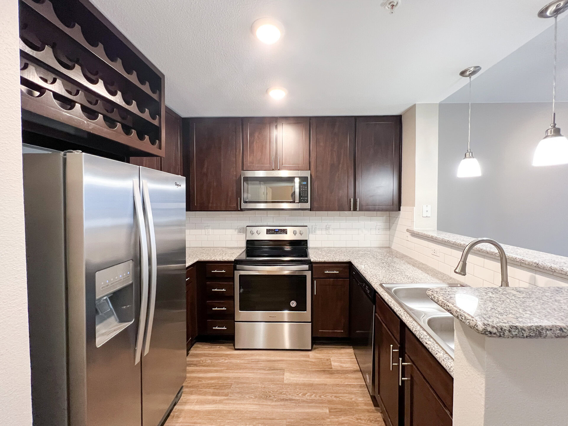 A modern kitchen featuring dark wood cabinetry and a light granite countertop. The stainless steel refrigerator is positioned on the left, next to a built-in microwave above a black stove. The backsplash consists of white subway tiles, and there are two pendant lights hanging above the countertop. A double sink is located on the right side, with a sleek faucet. The flooring appears to be a light wood laminate.