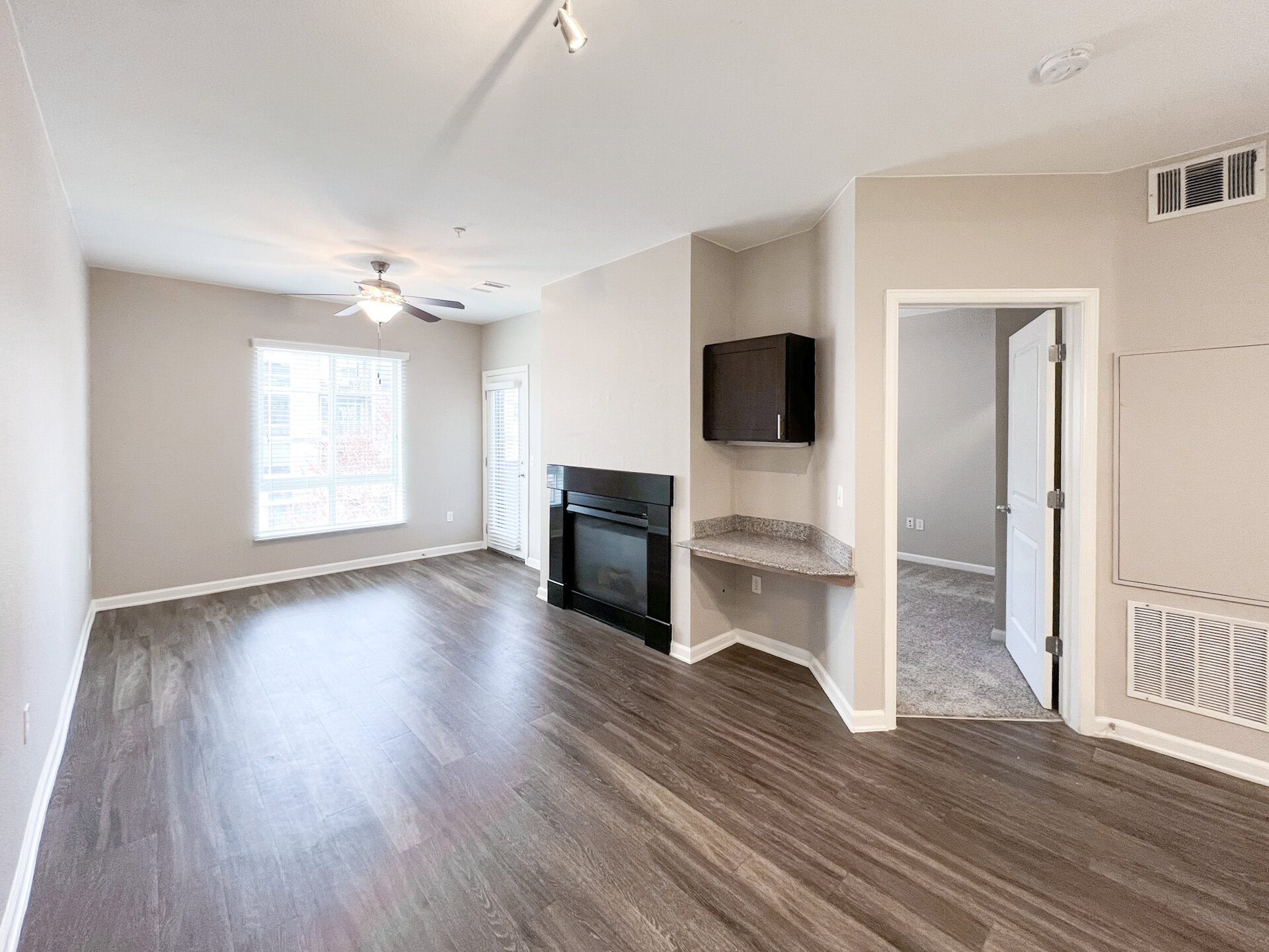 A spacious living area features a ceiling fan and a large window with blinds, allowing natural light to fill the room. The flooring is a dark wood laminate, and there is a black fireplace along one wall. Adjacent to the fireplace is a small countertop area with a granite surface and a dark cabinet above. A doorway leads to another room, while a door on the left opens to a small balcony or patio area. The walls are painted in a light neutral color, enhancing the bright and open feel of the space.