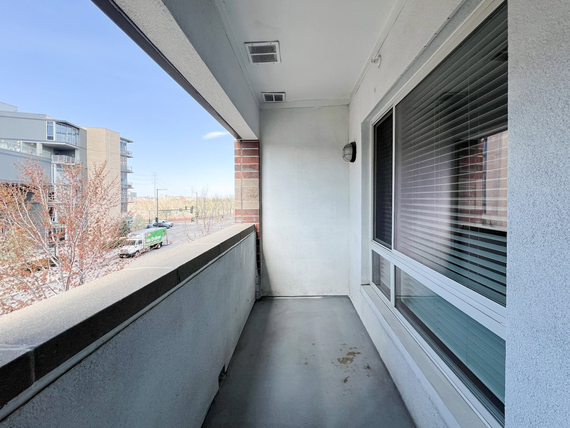 A large balcony with a concrete floor and a low wall. The wall has a smooth finish and a slight overhang. To the right, there are large windows with horizontal blinds. The view includes nearby buildings and a street with a green delivery truck parked. The sky is clear with a few clouds.
