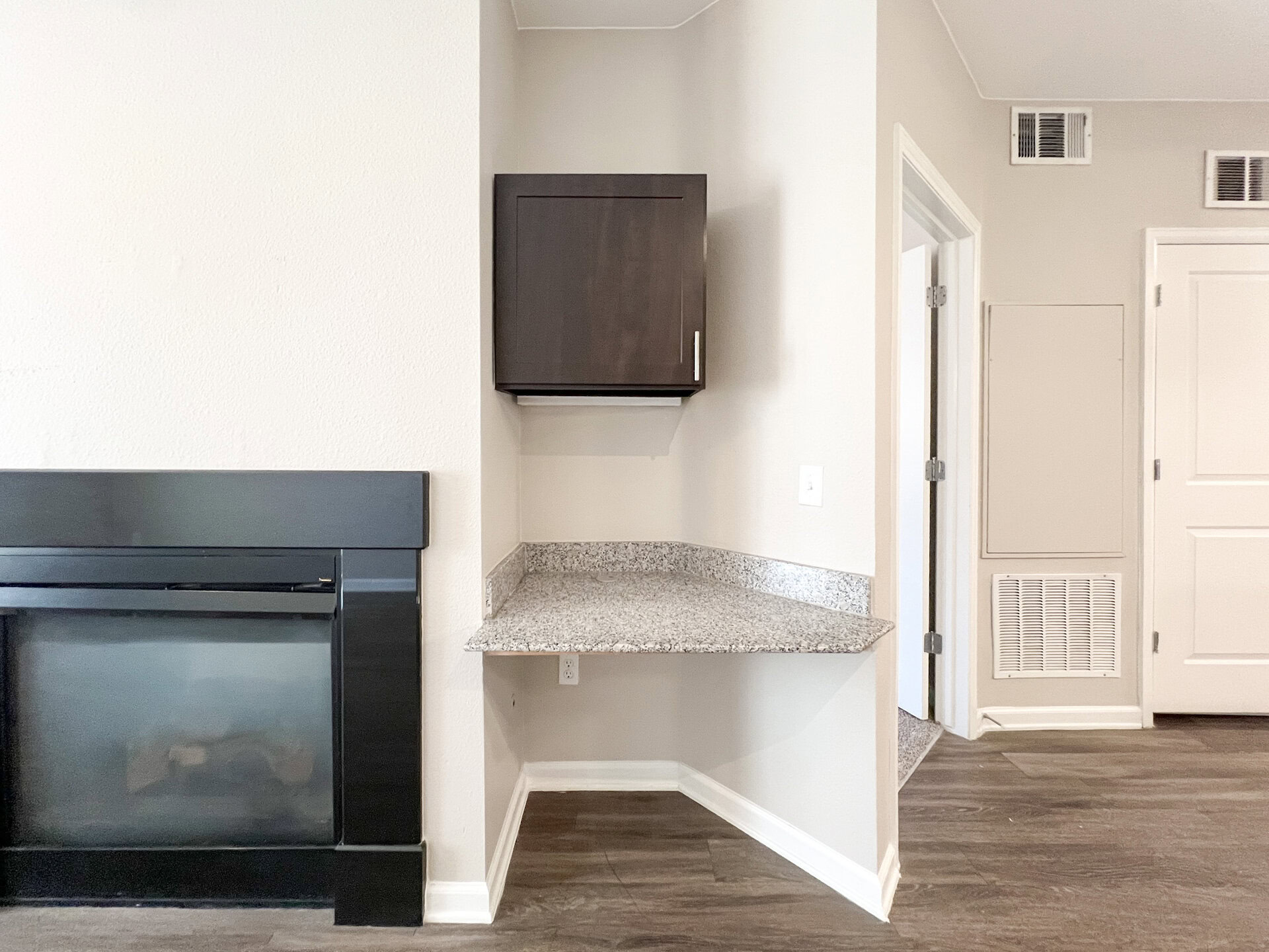 A corner of a room features a dark wood cabinet mounted on the wall above a light gray granite countertop. The countertop extends from the wall, providing a small workspace. To the right, there is a doorway leading to another room, and a vent is visible on the wall below the countertop. The flooring is dark wood, and the walls are painted a light color.