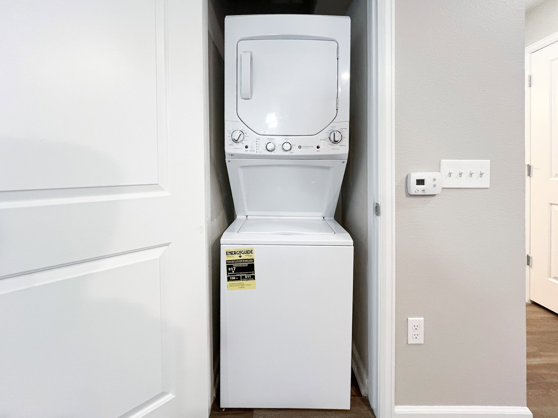 A stacked washer and dryer unit is positioned in a narrow space between two walls. The washer is at the bottom, featuring a top-loading design, while the dryer is mounted above it with a front-loading door. The unit is white and has control knobs and settings visible on the front. To the right, there is a wall with light switches and a thermostat, and a door is partially visible on the far right. The walls are painted a light gray color, and the flooring appears to be dark wood.