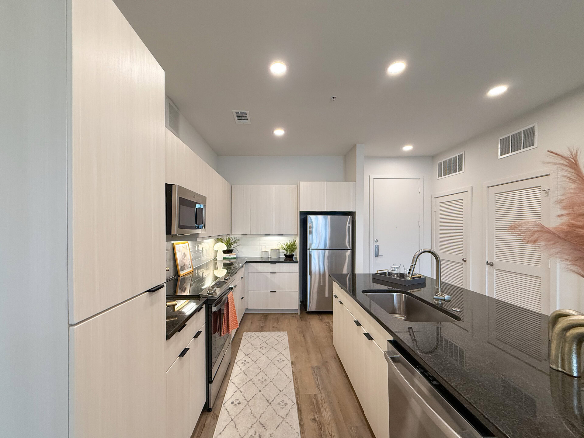A modern kitchen featuring light wood cabinetry and a sleek black countertop. The kitchen includes stainless steel appliances, including a refrigerator and a microwave. The flooring is a warm wood tone. Drawers are soft-closing with over 25 cabinets/drawers in the kitchen.