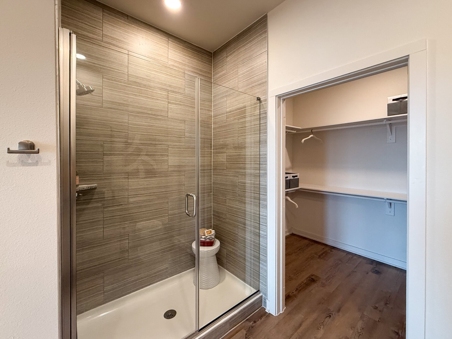 A modern standing shower features a glass-enclosed shower with a sleek, tiled wall in neutral tones. The flooring is a warm wood finish, adding a cozy touch to the space.