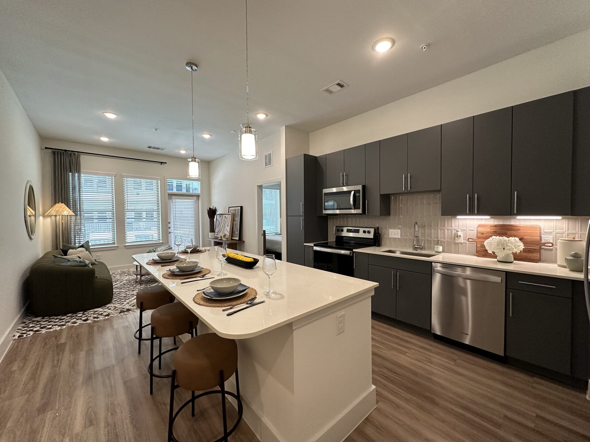 A modern kitchen and living area featuring an open layout. The kitchen has dark cabinetry, stainless steel appliances, and a light-colored countertop. A large island with seating for four is set with plates, glasses, and utensils. The living area includes a green sofa adorned with cushions, a decorative lamp, and a textured rug. Large windows with blinds allow natural light to fill the space, and a door leads outside. The overall decor is contemporary and inviting.