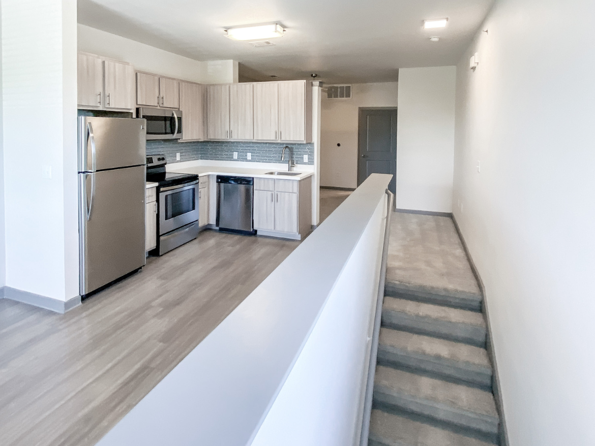 A modern kitchen features light wood cabinetry, stainless steel appliances including a refrigerator, oven, and dishwasher, and a white countertop. The backsplash is a subtle blue tile. The open layout leads to a carpeted staircase with a gray railing, transitioning to a hallway with a closed door at the end. The flooring is a light wood laminate, enhancing the contemporary feel of the space.