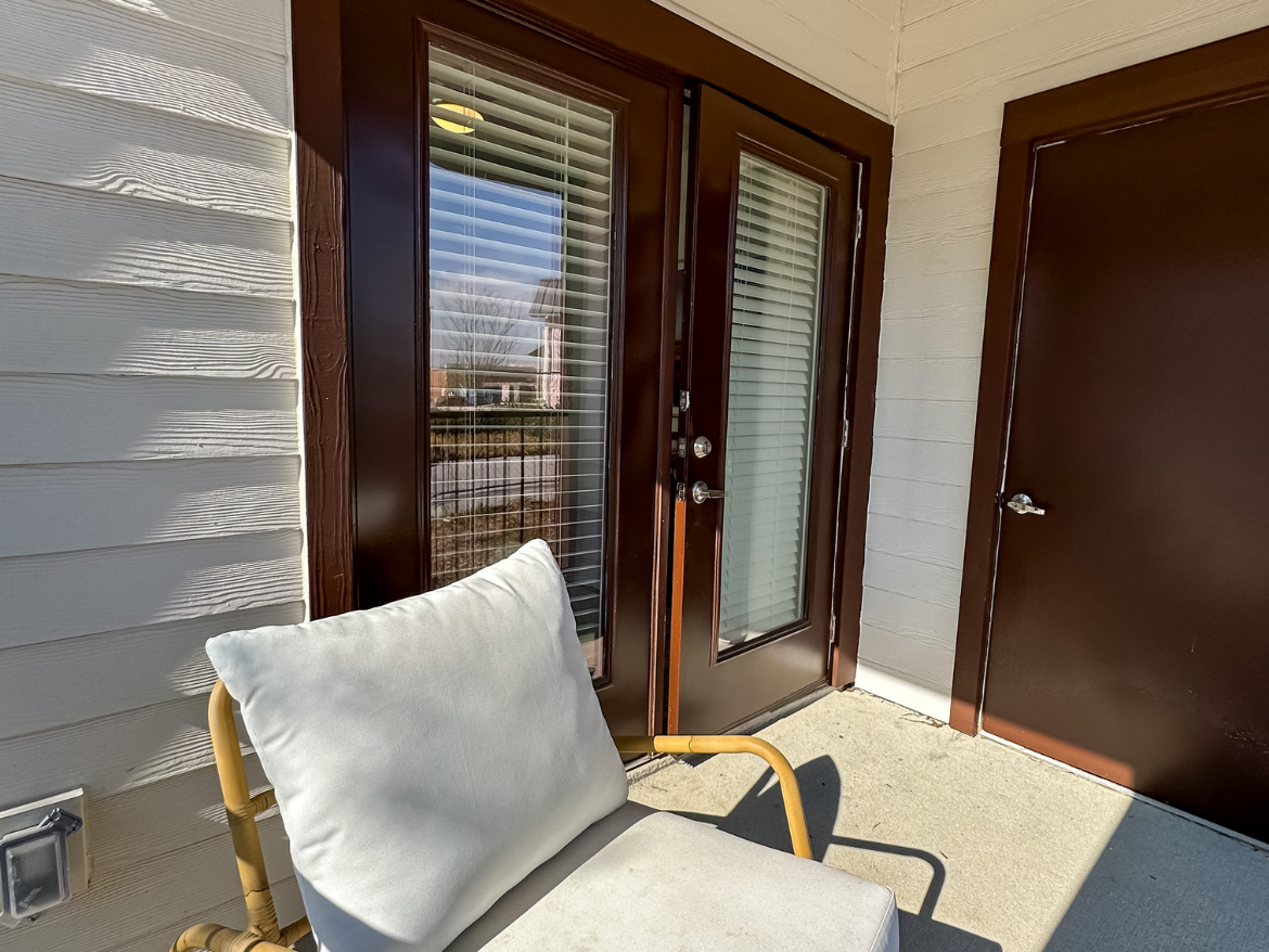 A small outdoor area features a light-colored cushion resting on a yellow metal chair. Two brown doors with glass panels are visible, one leading to the interior and the other likely a storage or utility door. The walls are white with a textured finish, and sunlight casts shadows across the surface. In the background, a glimpse of greenery and a pathway can be seen through the glass doors.