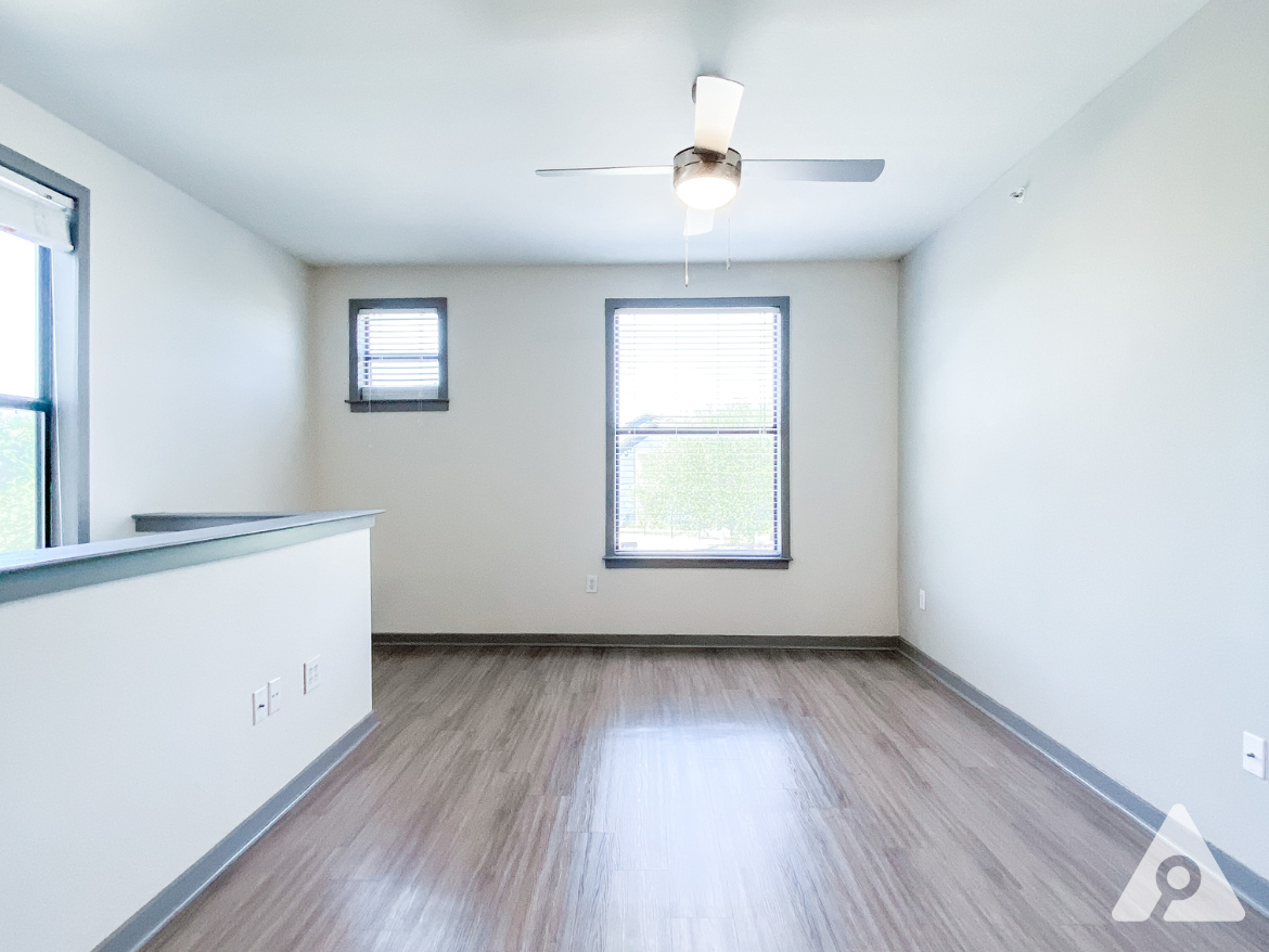 A spacious room features light-colored walls and a smooth, wooden floor. Two windows with blinds allow natural light to enter, and a ceiling fan is mounted in the center of the room. A low partition or ledge runs along one side, adding a subtle division to the space. The overall ambiance is bright and airy, suitable for various uses.