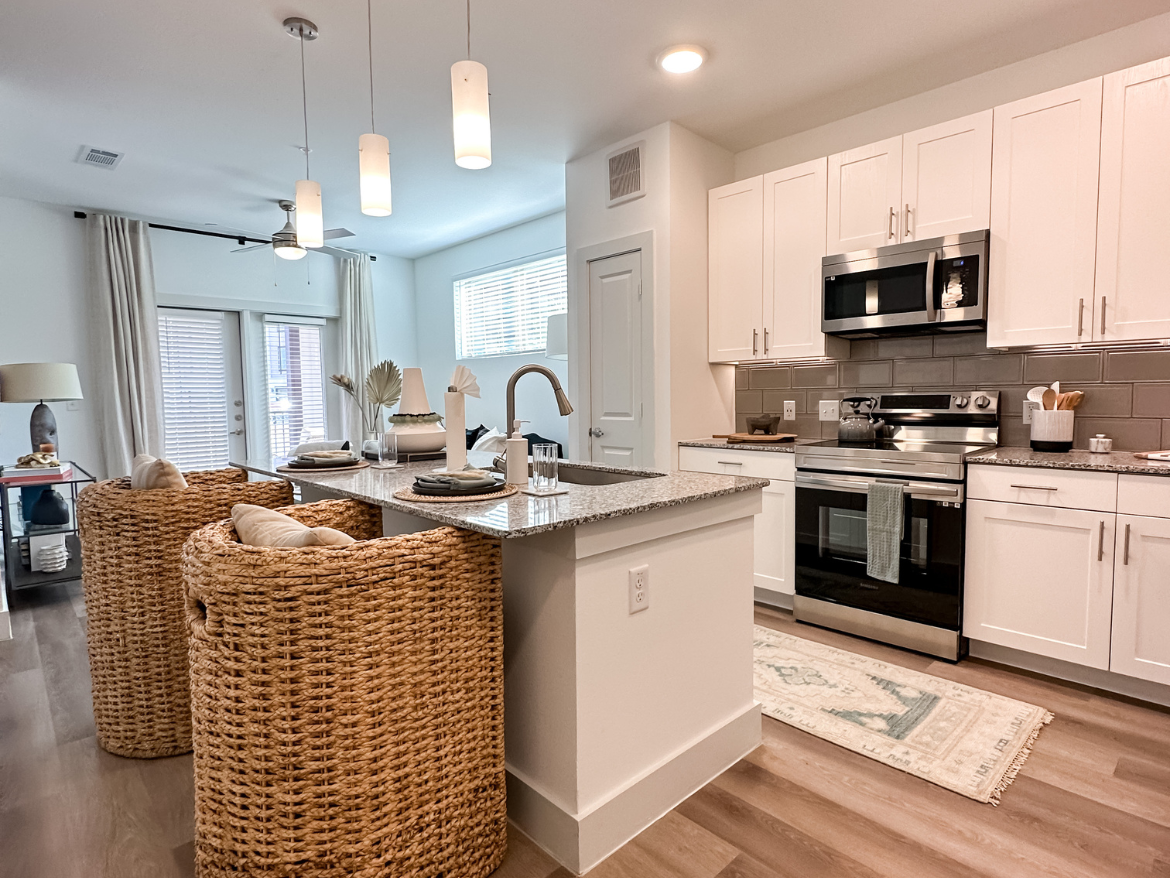 A modern kitchen features white cabinetry and stainless steel appliances, including a microwave and oven. The countertop is made of granite, with a stylish sink and faucet. Two woven bar stools with cushions are positioned at the island. Natural light streams in through a window, and there are decorative elements like a vase and a lamp on a nearby table. The flooring is a light wood, and a patterned rug adds warmth to the space. Pendant lights hang from the ceiling, enhancing the contemporary feel.