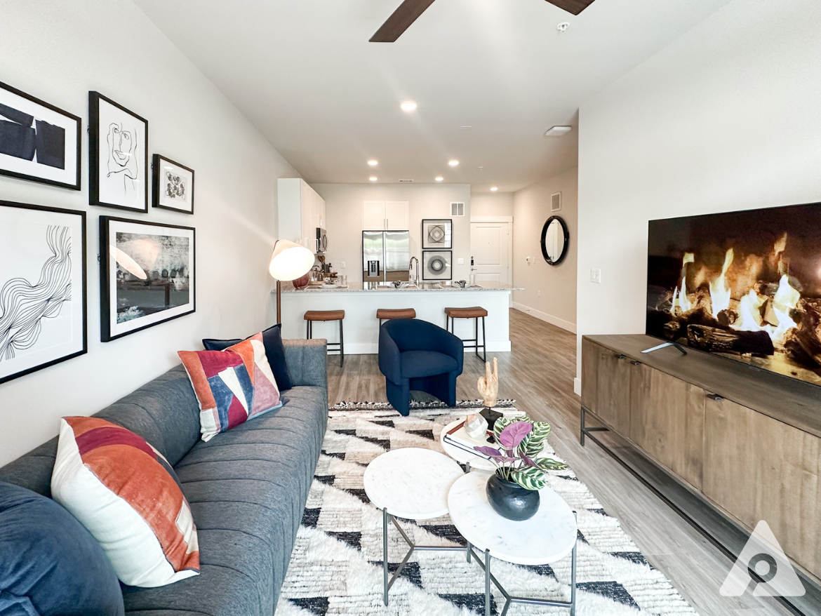 A modern living room features a gray sectional sofa adorned with colorful throw pillows. A round coffee table with a marble top sits on a patterned rug. Nearby, a dark wooden TV stand holds a large television displaying a fireplace scene. In the background, a kitchen area with white cabinetry and bar stools is visible, along with framed artwork on the walls and a floor lamp providing light. The space has a bright, airy feel with light wood flooring.