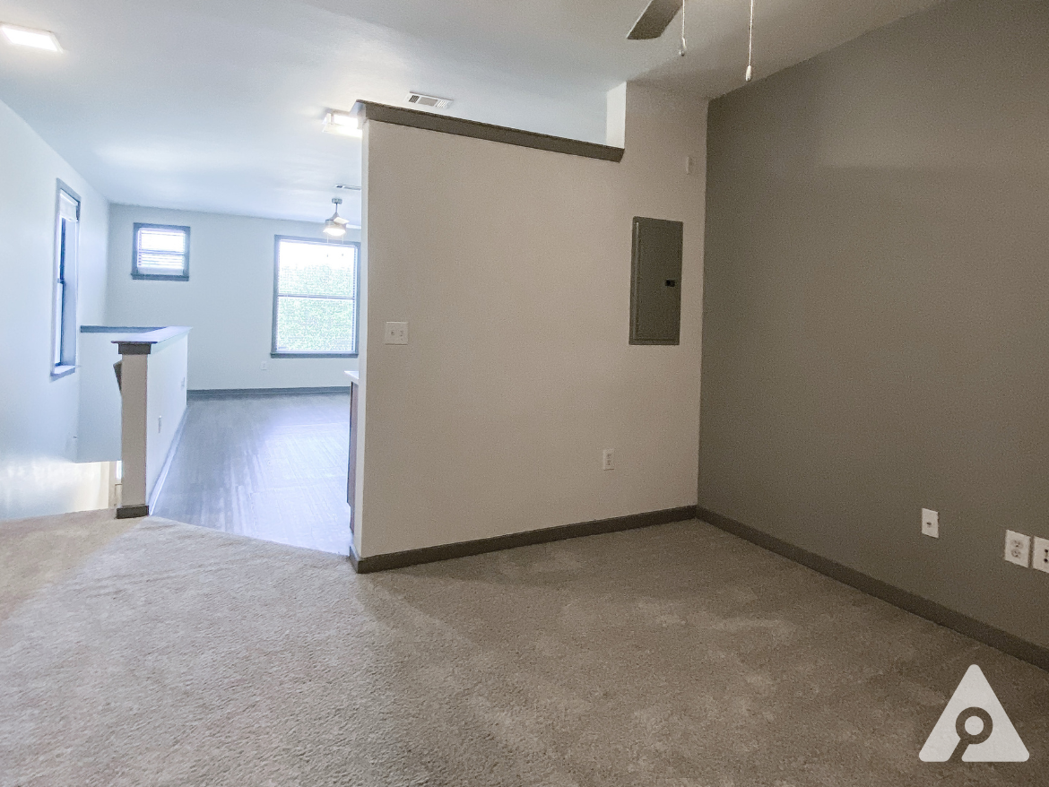 A spacious living area features light-colored walls and a carpeted floor. To the right, a gray wall with an electrical panel is visible. The room is illuminated by recessed lighting and a ceiling fan. Large windows allow natural light to enter, with one window framed by a view of greenery outside. The flooring transitions to a darker material, indicating a separation between areas.