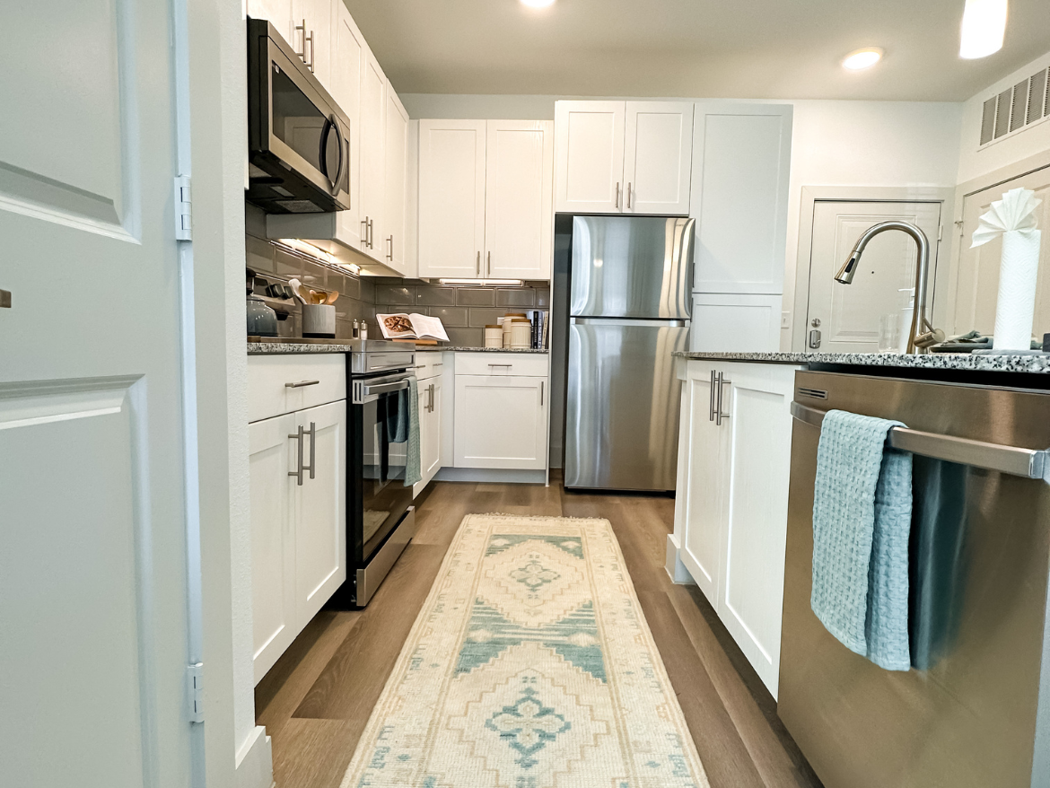 A modern kitchen featuring white cabinetry and stainless steel appliances. The space includes a microwave, oven, and refrigerator, with a granite countertop and a stylish backsplash. A light-colored area rug with a geometric pattern runs along the floor. The sink area has a sleek faucet, and a dish towel hangs from the dishwasher. Natural light illuminates the room, enhancing the clean and contemporary design.