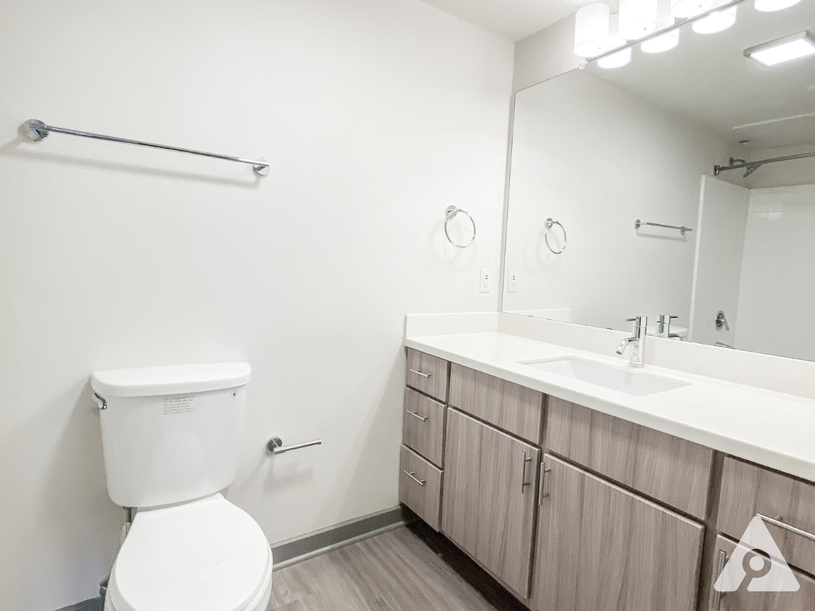 A modern bathroom featuring a white toilet and a sleek vanity with wooden cabinetry. The countertop is white and includes a rectangular sink with a chrome faucet. Above the sink, there is a large mirror and a light fixture with multiple bulbs. A towel bar is mounted on the wall, and the flooring is a light wood-like material. The shower area is visible in the background, enclosed with a glass door. The overall color scheme is neutral and bright, creating a clean and spacious feel.