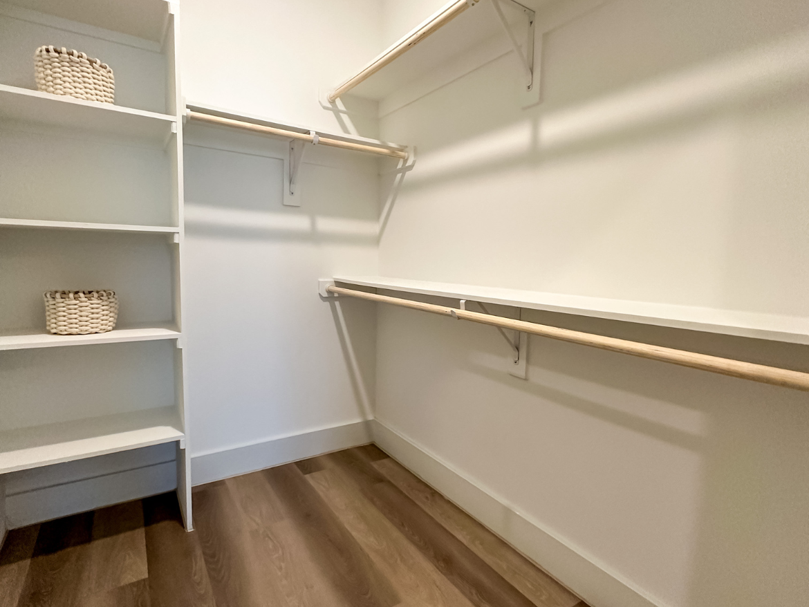 A spacious closet features white shelving units on the left side, with two woven baskets placed on the top shelf. On the right side, there are two wooden hanging rods supported by wall brackets, providing ample space for hanging clothes. The floor is covered with light-colored wood, and the walls are painted white, creating a bright and clean appearance.