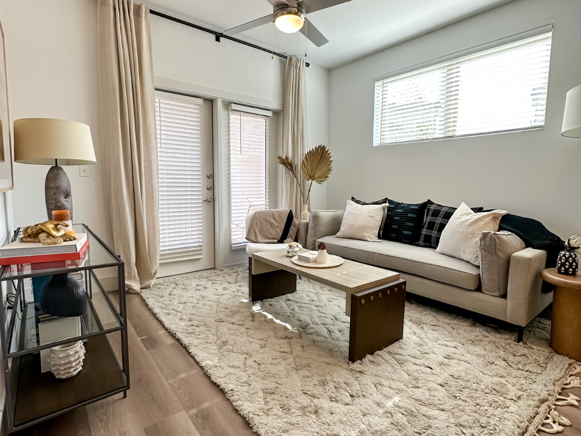 A cozy living room features a light-colored sofa adorned with various patterned and solid throw pillows. A wooden coffee table with a unique design sits in front of the sofa, topped with decorative items. The floor is covered with a plush, textured rug. To the side, a glass console table displays decorative objects and books. Natural light streams in through large windows with blinds, and soft curtains frame the space. A ceiling fan adds to the room's comfort, while a small side table with a lamp completes the inviting atmosphere.