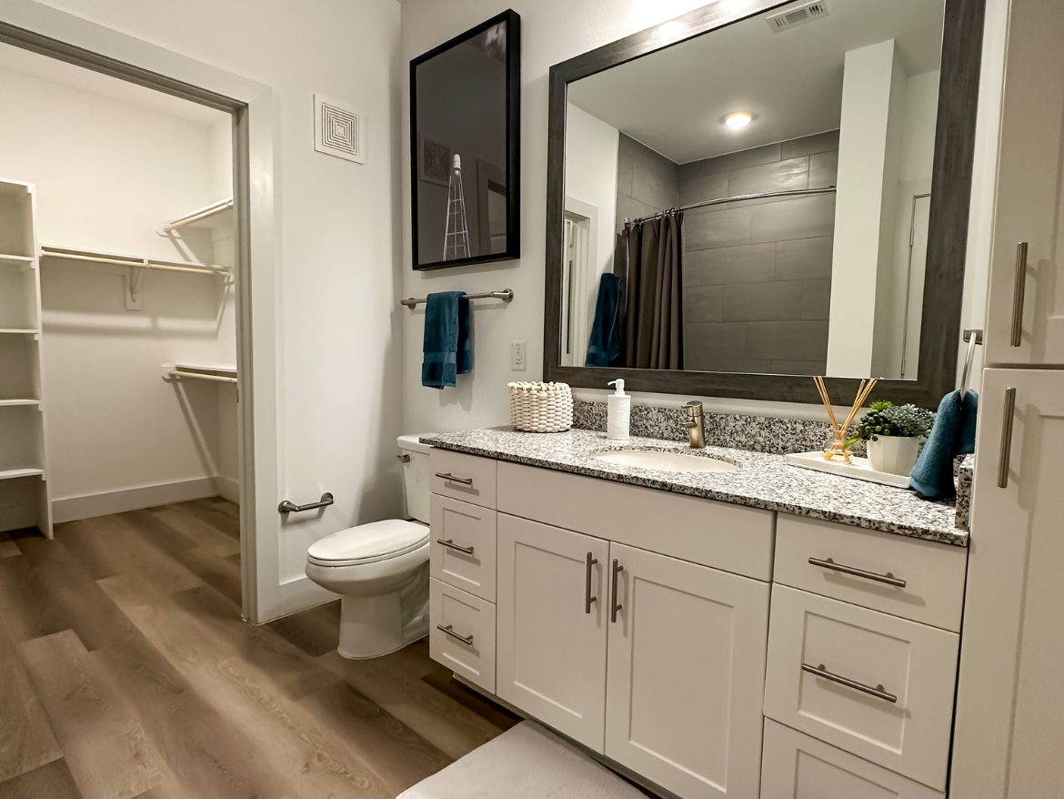 A modern bathroom features a double vanity with a granite countertop, equipped with a sink and stylish faucet. Above the vanity, a large rectangular mirror is framed in dark wood. The walls are painted white, and there is a gray tiled section behind the shower area, which is enclosed by a curtain. A small potted plant and decorative items are placed on the countertop. To the left, an open closet with shelving provides additional storage space. The flooring is a light wood laminate, enhancing the contemporary feel of the space.