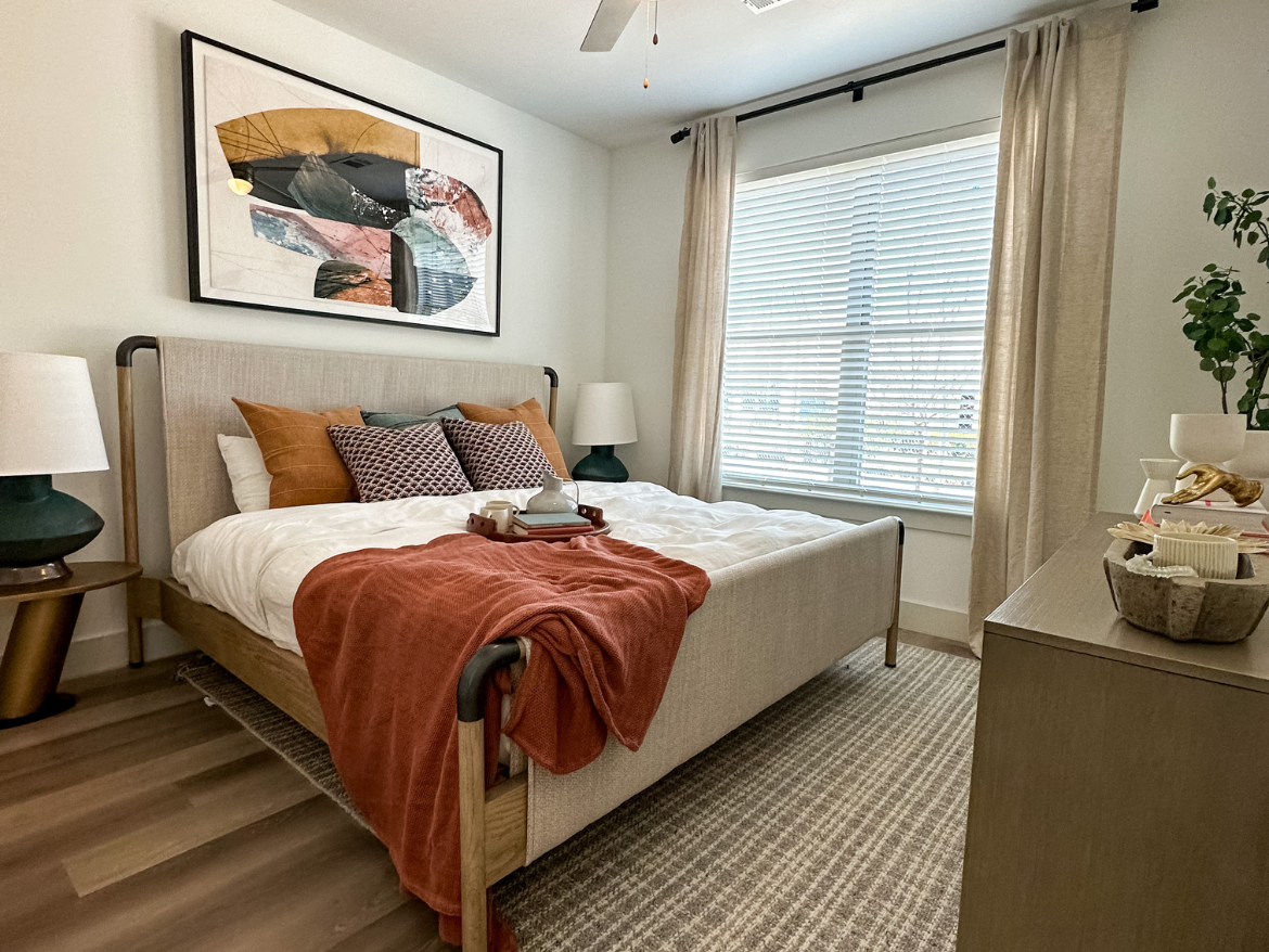 A cozy bedroom features a light-colored bed with a textured headboard, adorned with orange and patterned throw pillows. A soft, rust-colored blanket drapes over the side. Two bedside lamps with green bases sit on either side of the bed. Above the bed, an abstract artwork adds a pop of color. Natural light filters through a window with white blinds, flanked by beige curtains. A small nightstand holds decorative items, including a bowl and a plant, while a textured area rug covers the floor.