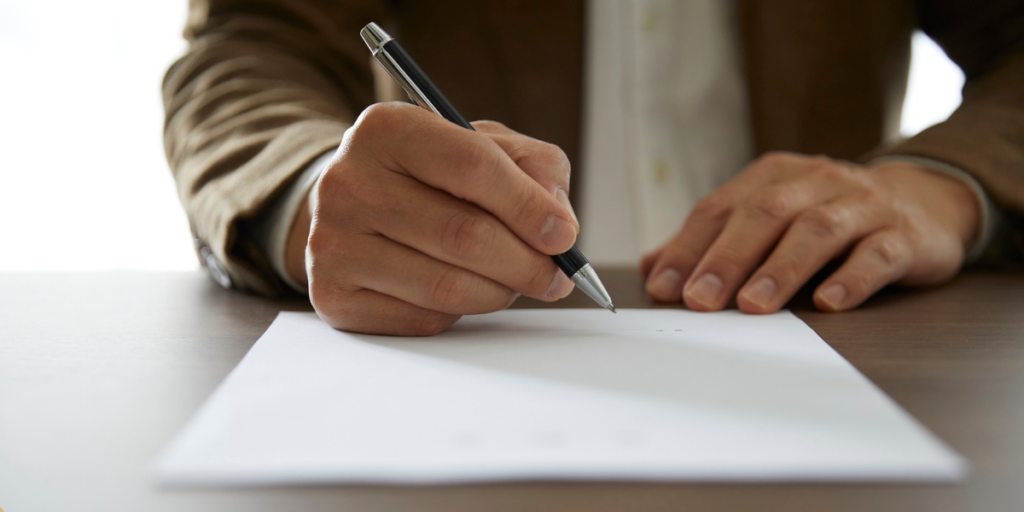 A hand is holding a pen, poised to write on a blank sheet of paper placed on a wooden table. The person appears to be wearing a brown blazer over a white shirt. The background is softly blurred, suggesting a well-lit indoor setting. Should you renew your lease or move?