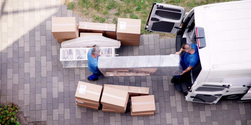 Movers are unloading a large, wrapped item from the back of a white delivery van. They are positioned on a paved area, surrounded by several cardboard boxes stacked nearby. The boxes are sealed and labeled, indicating they contain various items. The scene suggests a moving or delivery process, with one person lifting the large item while the other assists. The background features a patch of grass, adding a touch of greenery to the setting.