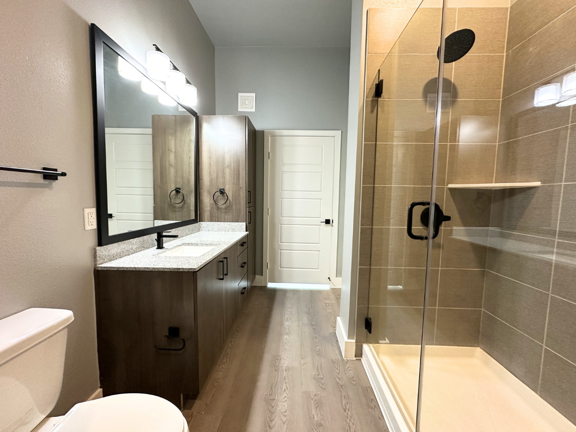 A modern bathroom features a double vanity with a granite countertop, two sinks, and a large rectangular mirror framed in black. The cabinetry is dark wood, providing storage space. To the left, a white toilet is positioned next to a towel bar. On the right, a glass-enclosed shower showcases a tiled interior with a built-in shelf and a black showerhead. The walls are painted a soft gray, and the flooring is a light wood laminate. A white door leads to another area of the home.