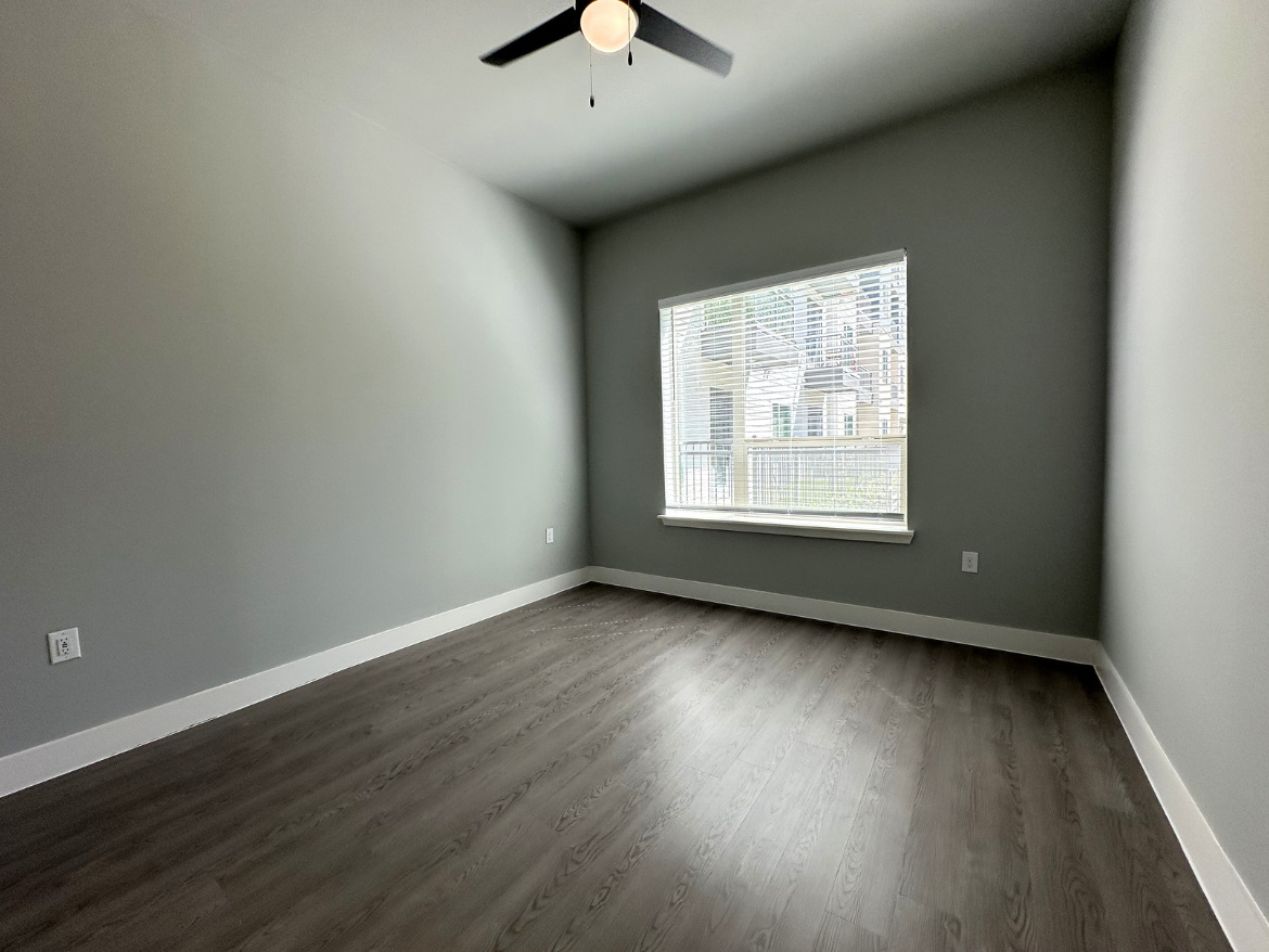 A spacious room with light gray walls and a smooth, dark wood floor. A ceiling fan with black blades hangs from the ceiling. Natural light streams in through a large window with white blinds, offering a view of a balcony and nearby buildings. The room is empty, creating a clean and open atmosphere.