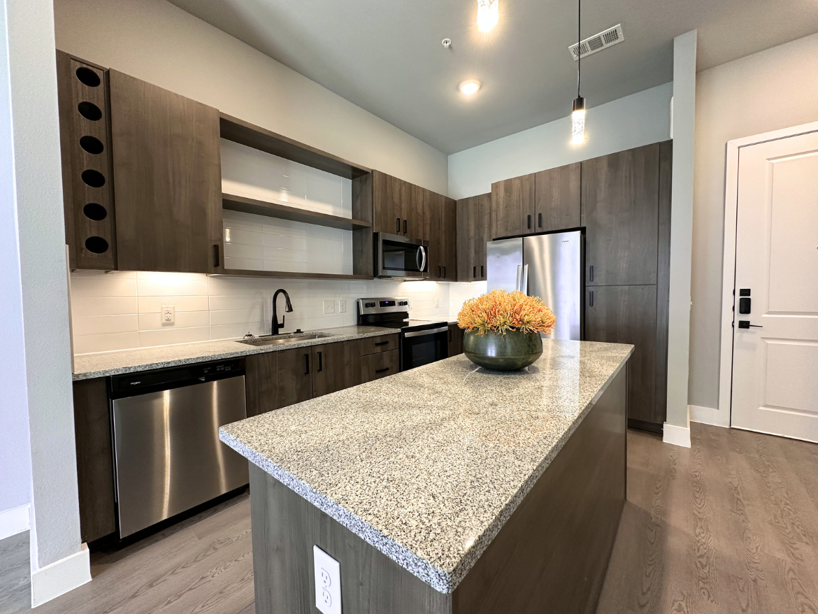 A modern kitchen features sleek, dark wood cabinetry with a mix of closed and open shelves. The countertop is a light-colored granite, complementing the stainless steel appliances, including a refrigerator, oven, and dishwasher. A black faucet is installed above the sink, which is positioned against a white tiled backsplash. A decorative bowl filled with orange flowers sits on the countertop, adding a pop of color. The flooring is a light wood, and the entrance door is visible in the background.