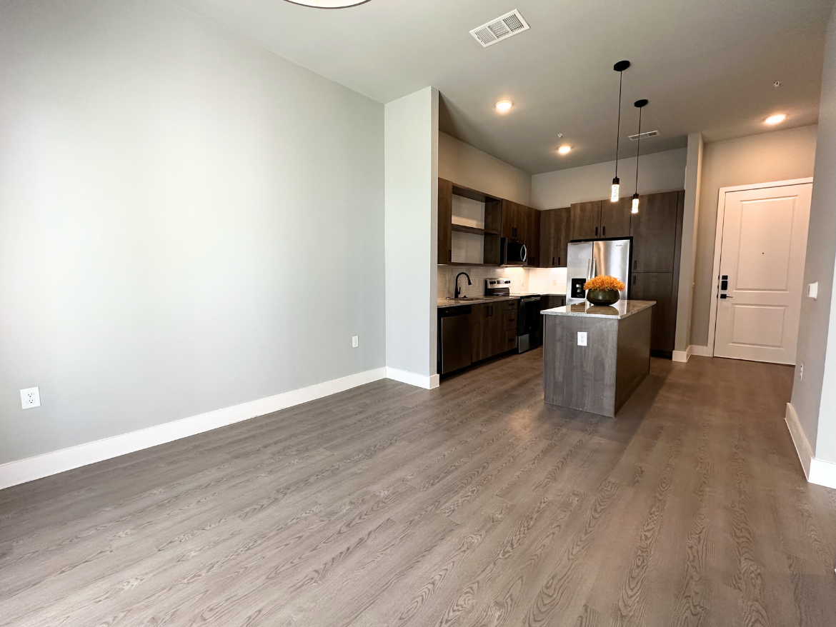 A modern, open-concept living space features a kitchen with dark wood cabinetry and stainless steel appliances, including a refrigerator and stove. A central island with a light-colored countertop serves as a focal point, adorned with a decorative vase of flowers. The flooring is a light wood laminate, and the walls are painted in a soft gray. Pendant lights hang above the island, and a front door is visible on the right side of the image. The overall ambiance is bright and contemporary.