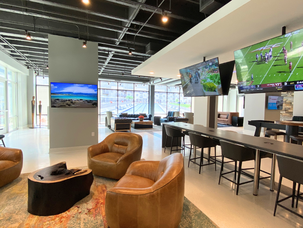 A modern lounge area features comfortable brown leather chairs arranged around a unique wooden coffee table. Two large flat-screen TVs are mounted on the walls, displaying a beach scene and a sports game. The space is well-lit with natural light streaming in through large windows, and there are additional seating options, including a long table with high-backed chairs. The decor includes a colorful area rug and some decorative sculptures in the background.