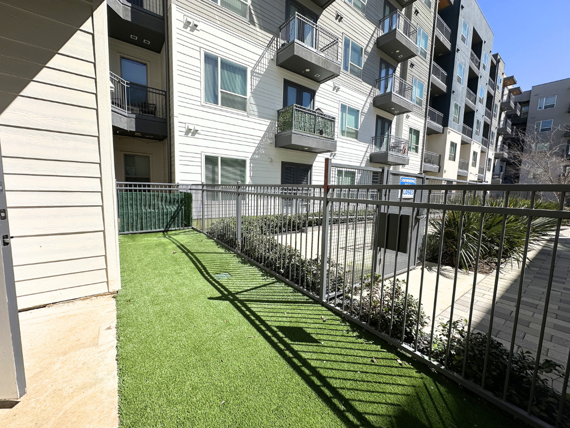 A narrow pathway lined with artificial grass leads to a fenced area adjacent to a multi-story apartment building. The pathway is bordered by a metal railing, and there are several balconies visible on the building's facade. In the background, a sign indicates that the swimming pool area is closed. The scene is bright, with clear blue skies and sunlight casting shadows on the ground. Lush greenery is present along the pathway and in the surrounding area.