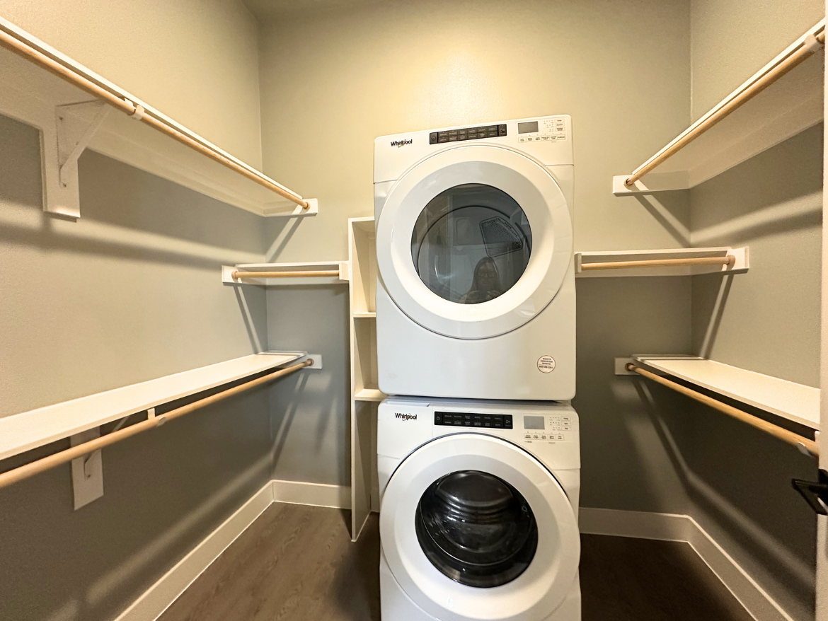 A compact laundry area features a stacked washer and dryer in white. The appliances are positioned against a light gray wall, with several wooden rods and shelves installed around them for storage and drying. The flooring is a dark wood laminate, adding contrast to the space. The overall design is functional and modern, maximizing the use of the available area.