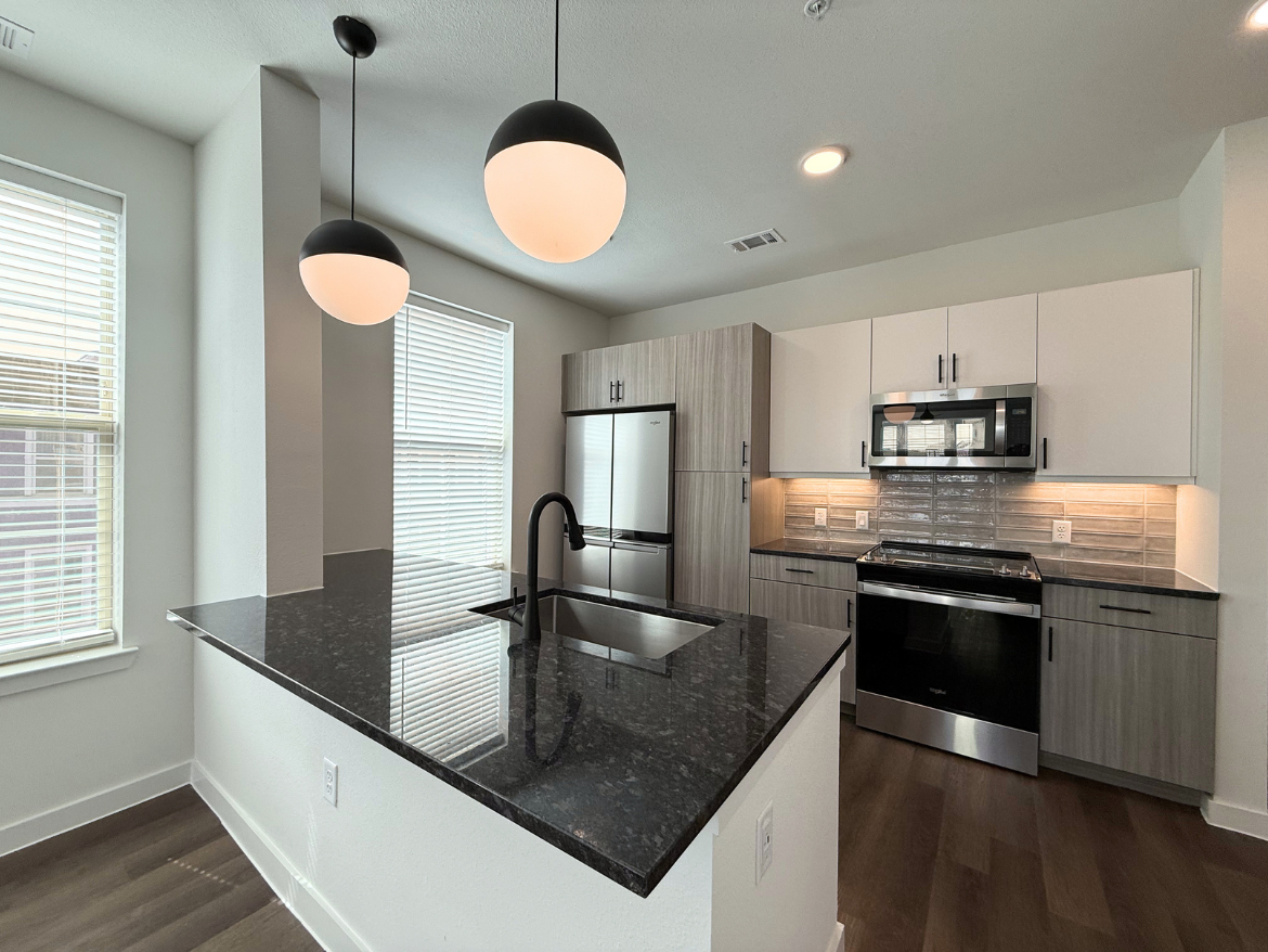 A modern kitchen featuring a large island with a dark granite countertop and a sleek black faucet. Above the island, two round pendant lights provide illumination. The kitchen has a combination of light and dark cabinetry, with stainless steel appliances including a refrigerator and a microwave. The backsplash consists of textured gray tiles, and there are large windows with blinds allowing natural light to enter the space. The flooring is a warm wood tone, enhancing the contemporary feel of the kitchen.
