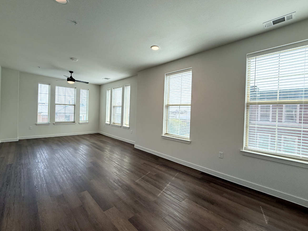 A spacious, well-lit room features large windows with blinds, allowing natural light to fill the space. The flooring is a rich, dark wood, enhancing the modern aesthetic. A ceiling fan is centrally located, and the walls are painted in a light color, contributing to the airy feel of the room. The layout is open, providing flexibility for furniture arrangement.