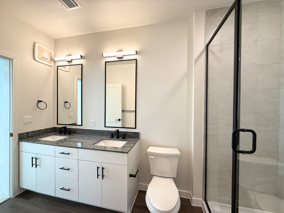 A modern bathroom features a double vanity with a dark granite countertop and two rectangular sinks. Above the sinks are two framed mirrors with black borders, flanked by wall-mounted light fixtures. The cabinetry is white with black handles. To the right, a glass shower enclosure with a black frame showcases light gray tiles. A white toilet is positioned next to the vanity, and a door leads to another space. The overall color scheme is neutral, with light walls and dark accents.