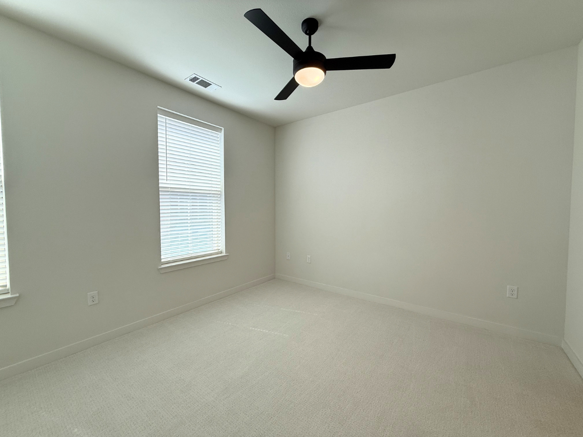 A bright, empty room with light-colored walls and a soft carpeted floor. Two windows with white blinds allow natural light to enter, illuminating the space. A modern black ceiling fan with a light fixture is mounted on the ceiling, adding a contemporary touch. The overall atmosphere is clean and minimalistic, ideal for various uses.