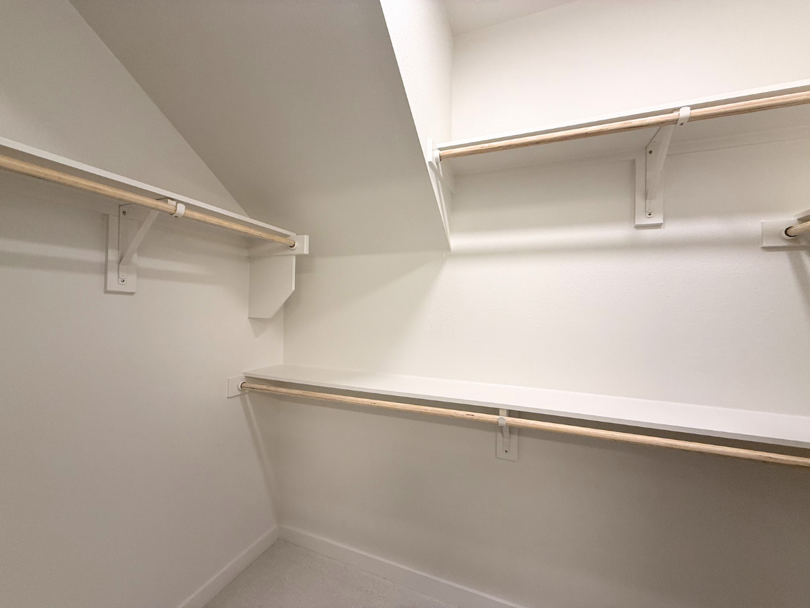 A small, organized closet features two wooden hanging rods supported by white brackets, positioned on opposite walls. There is a white shelf above the rods, and the walls and floor are painted in a clean, light color, creating a bright and spacious feel. The ceiling slopes gently, adding to the unique shape of the closet.