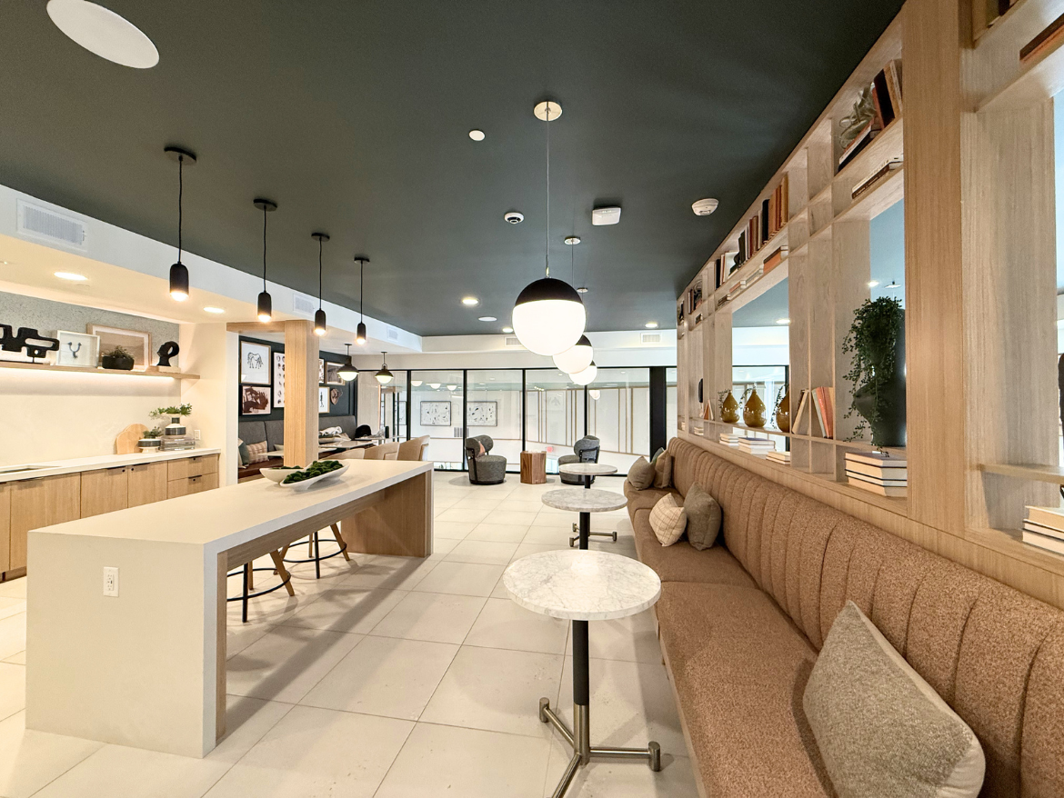 A modern interior space features a long, minimalist white countertop with wooden accents, complemented by bar stools. The area is well-lit with pendant lights hanging from a dark ceiling. To one side, a cozy seating arrangement includes a tufted bench with decorative pillows and small marble-topped tables. The walls are adorned with framed artwork and shelves displaying books and decorative items, creating a stylish and inviting atmosphere. Large windows allow natural light to fill the space, enhancing the overall ambiance.