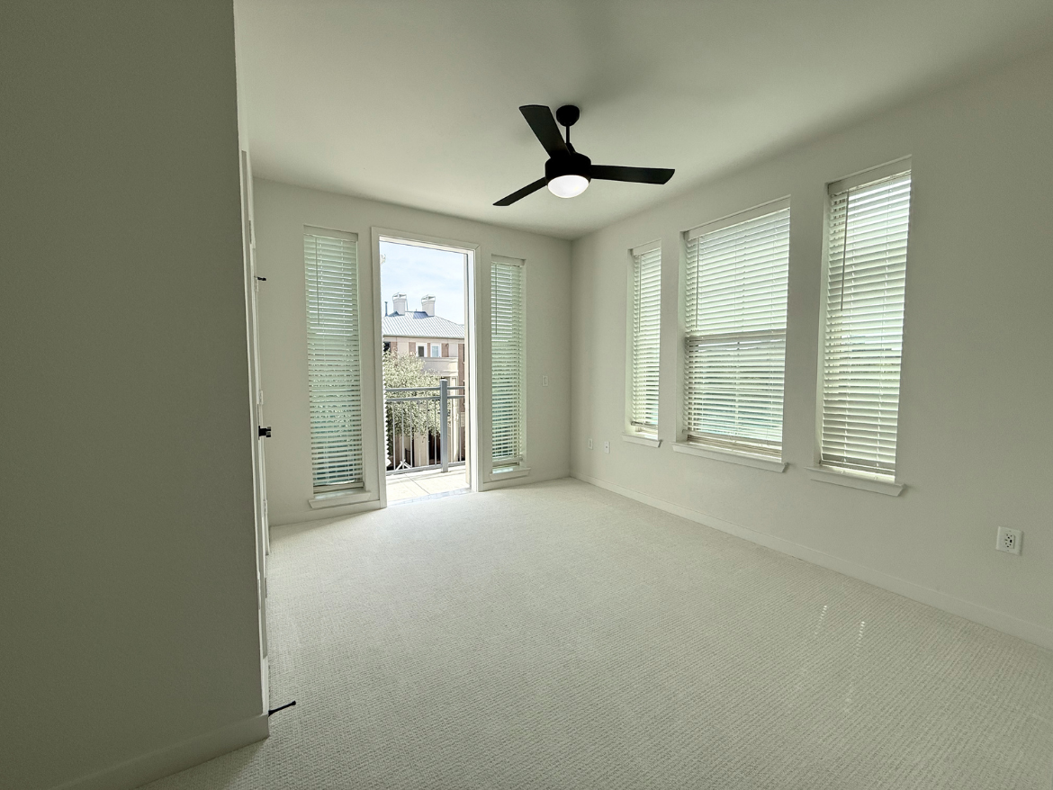 A bright, empty room features light-colored walls and a soft carpeted floor. Large windows with blinds allow natural light to fill the space, and a ceiling fan is mounted in the center. A door leads to a small balcony, visible through the open doorway. The overall atmosphere is clean and airy, with a minimalist design.
