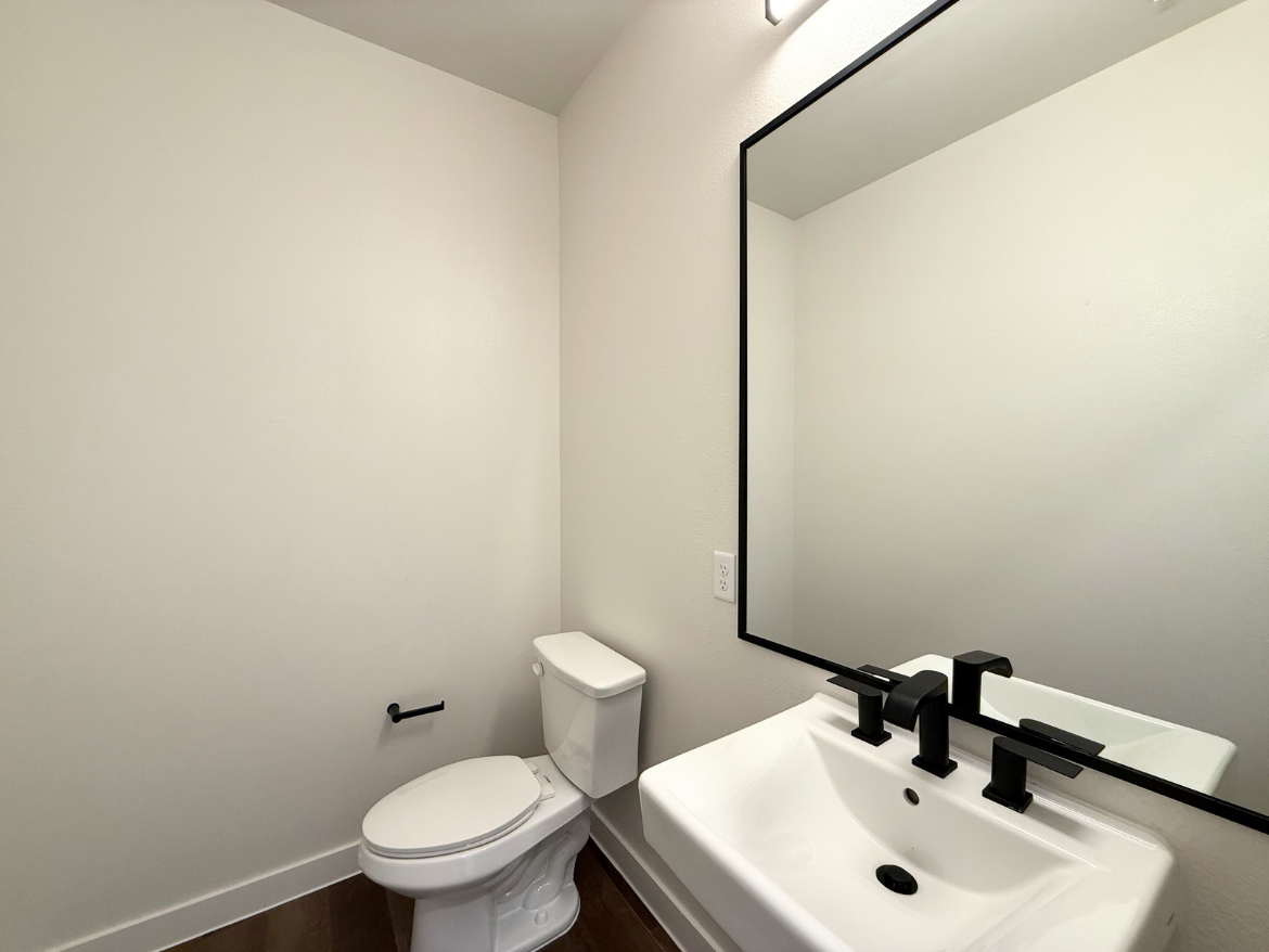 A modern bathroom features a white toilet and a sleek, white sink with black faucets. Above the sink is a large rectangular mirror framed in black. The walls are painted in a light color, and the flooring is a dark wood. A small black towel bar is mounted on the wall next to the toilet. The overall design is minimalist and contemporary.