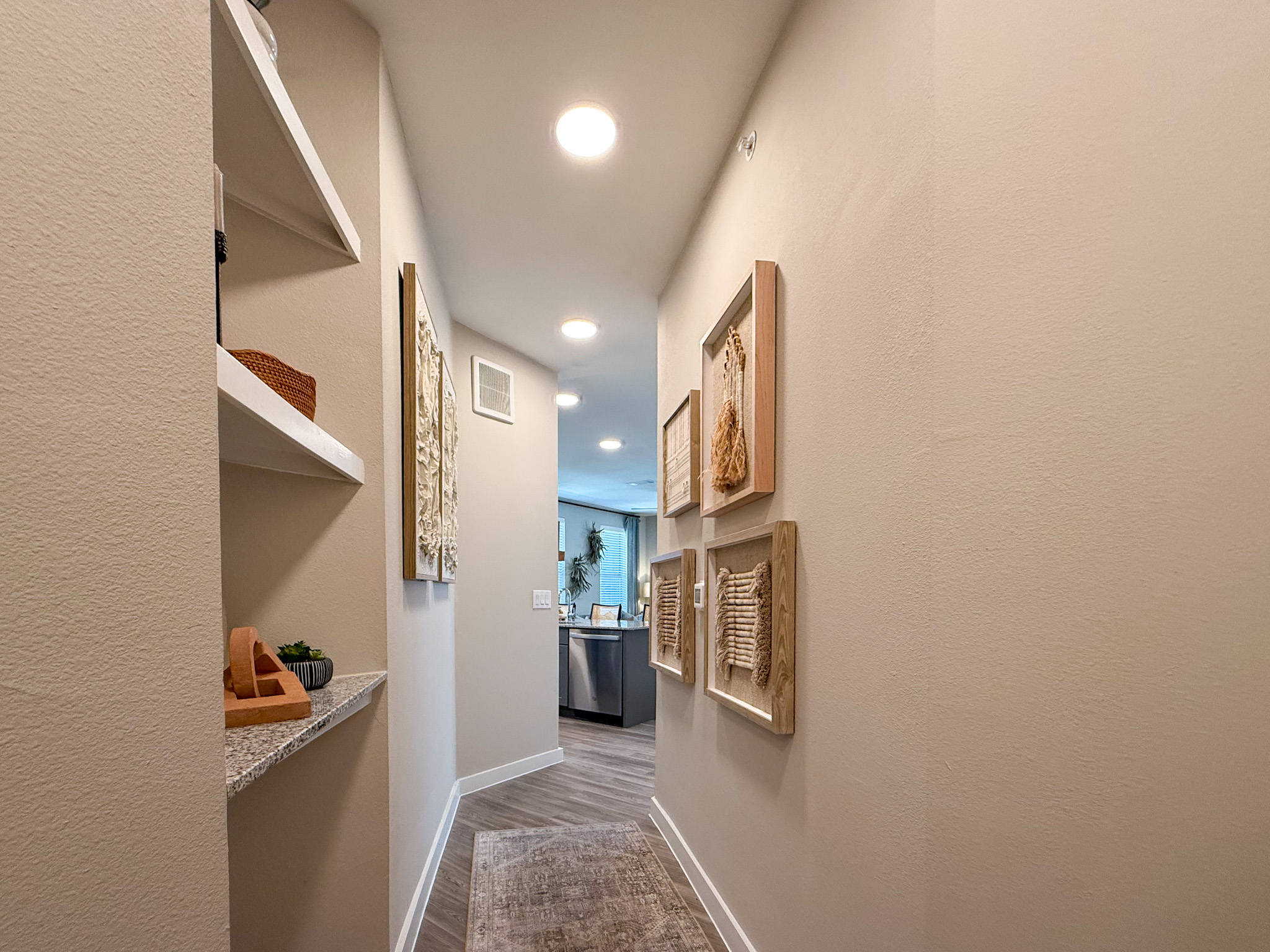 A narrow hallway features light-colored walls and recessed lighting. On the left, there is a floating shelf with a small decorative item and a potted plant. The right wall displays a series of framed wall art pieces with textured designs. At the end of the hallway, a glimpse of a kitchen is visible, showcasing modern appliances and a window with natural light filtering in. The flooring is a light wood finish, adding warmth to the space.