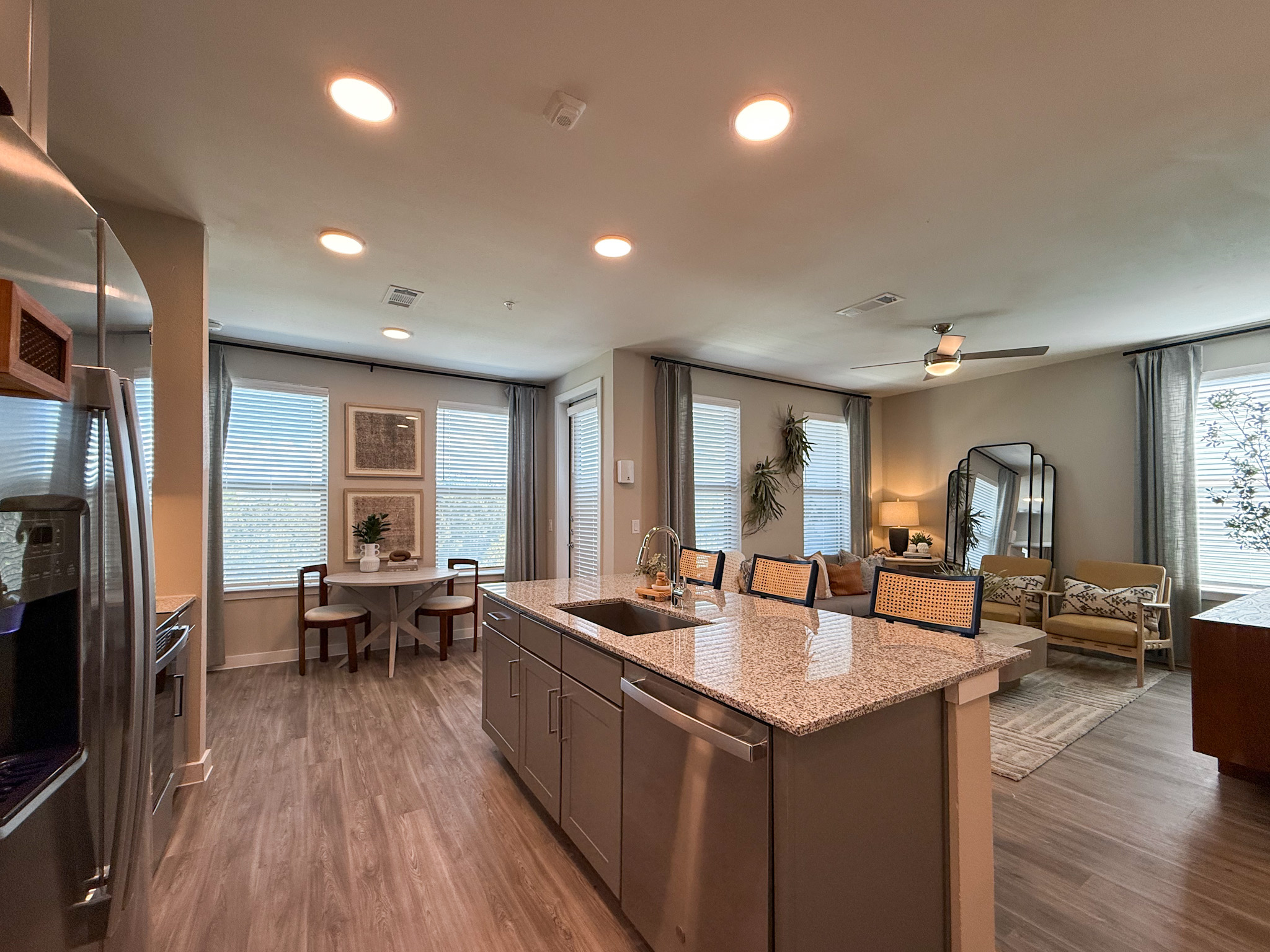 A cozy San Antonio apartment kitchen and living area featuring a large granite island with a sink and seating. Stainless steel appliances, including a refrigerator and dishwasher, are present. Natural light floods the space through multiple windows with blinds. A round dining table with two chairs is positioned near the windows, while the living area includes a cozy sofa, decorative pillows, and a stylish mirror.