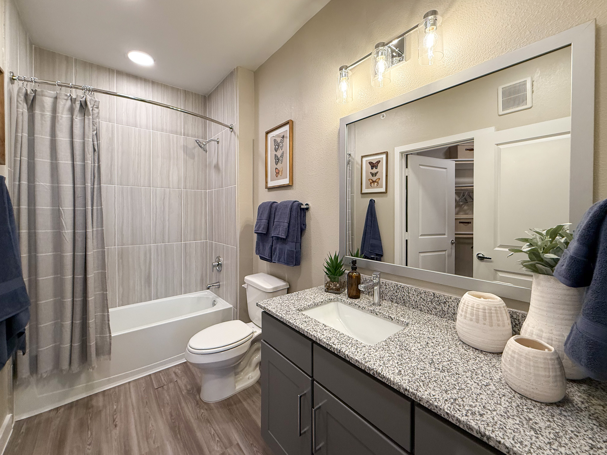 A modern San Antonio apartment bathroom features a bathtub with a curved shower rod and a gray striped curtain. The walls are tiled with light gray tiles. A white toilet is positioned next to the bathtub. The vanity has a granite countertop with a white sink and a sleek faucet. The lighting is provided by stylish glass fixtures.