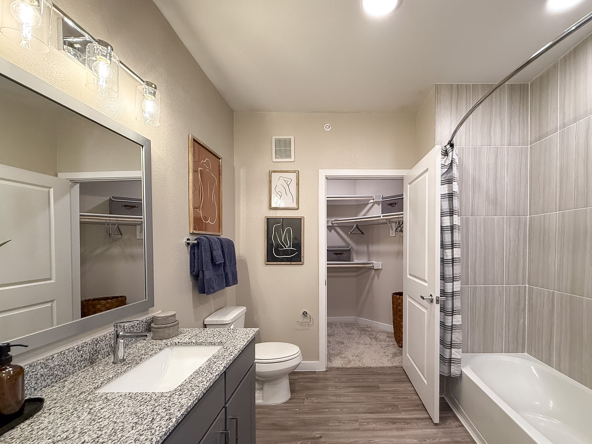 A modern San Antonio apartment bathroom features a bathtub with a curved shower rod and a gray striped curtain. The walls are tiled with light gray tiles. A white toilet is positioned next to the bathtub. The vanity has a granite countertop with a white sink and a sleek faucet. The lighting is provided by stylish glass fixtures.