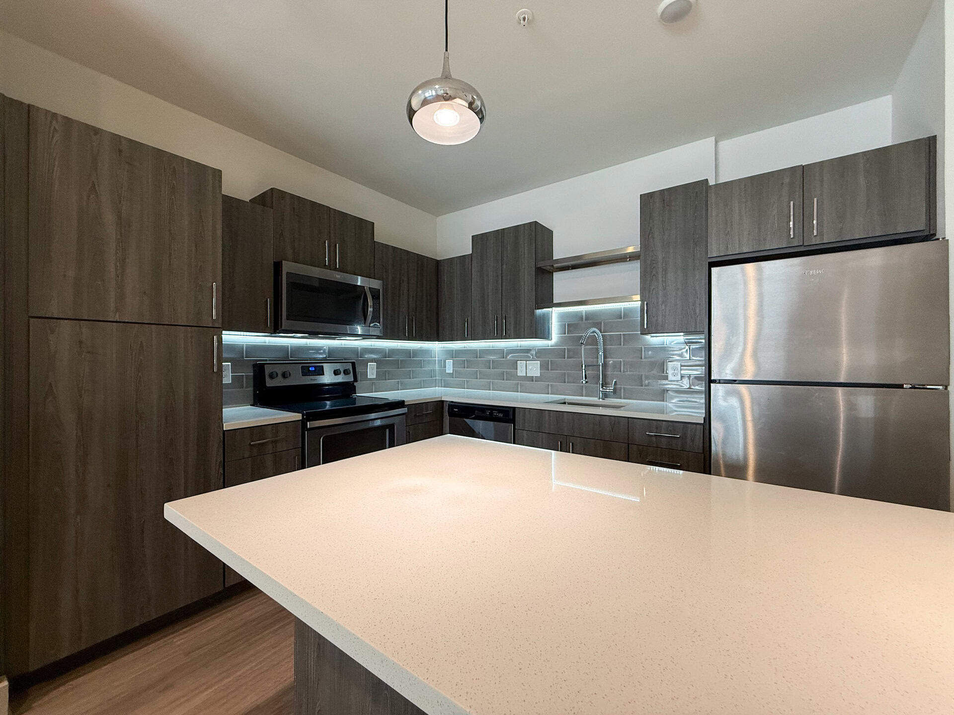 A modern kitchen featuring dark wood cabinetry and stainless steel appliances. A sleek countertop with a light-colored finish extends into an island, providing additional workspace. The backsplash is composed of gray tiles, and under-cabinet lighting enhances the overall ambiance.