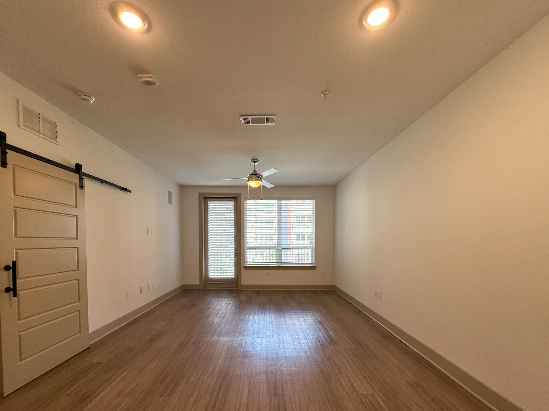 A spacious living room features a ceiling fan with a light fixture hangs from the ceiling. On one side, there is a sliding barn door leading to another area. A large window with horizontal blinds allows natural light to enter, and a glass door opens to a balcony.