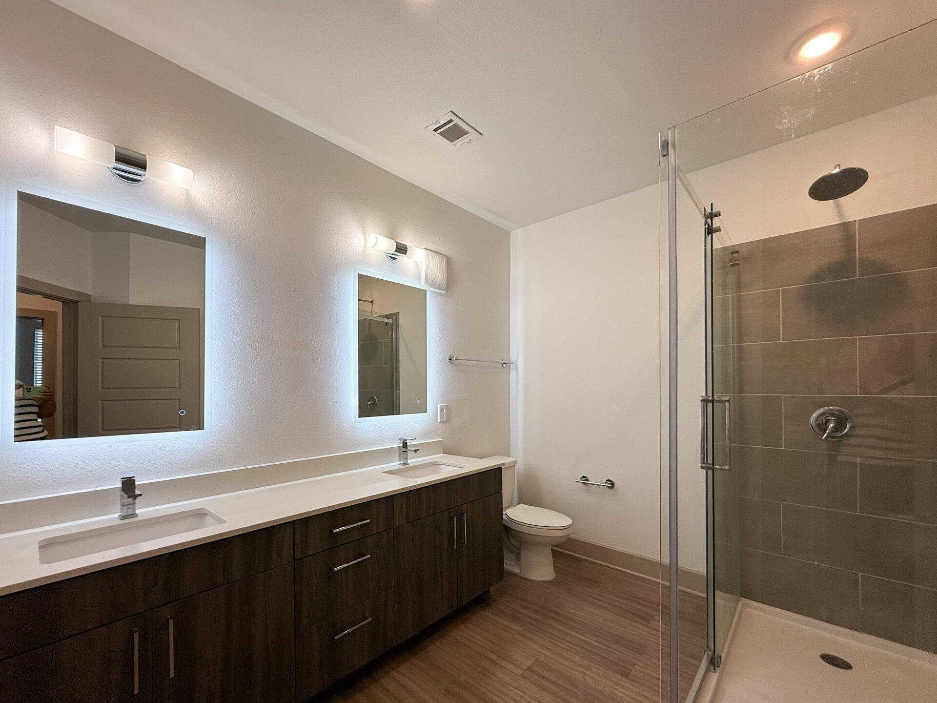 A modern bathroom features a double vanity with a sleek white countertop and dark wood cabinetry. Above the vanity, two illuminated mirrors are mounted on the wall. To the right, a glass shower enclosure showcases a tiled interior with a rainfall showerhead. The overall design is clean and contemporary, with neutral tones and ample lighting.